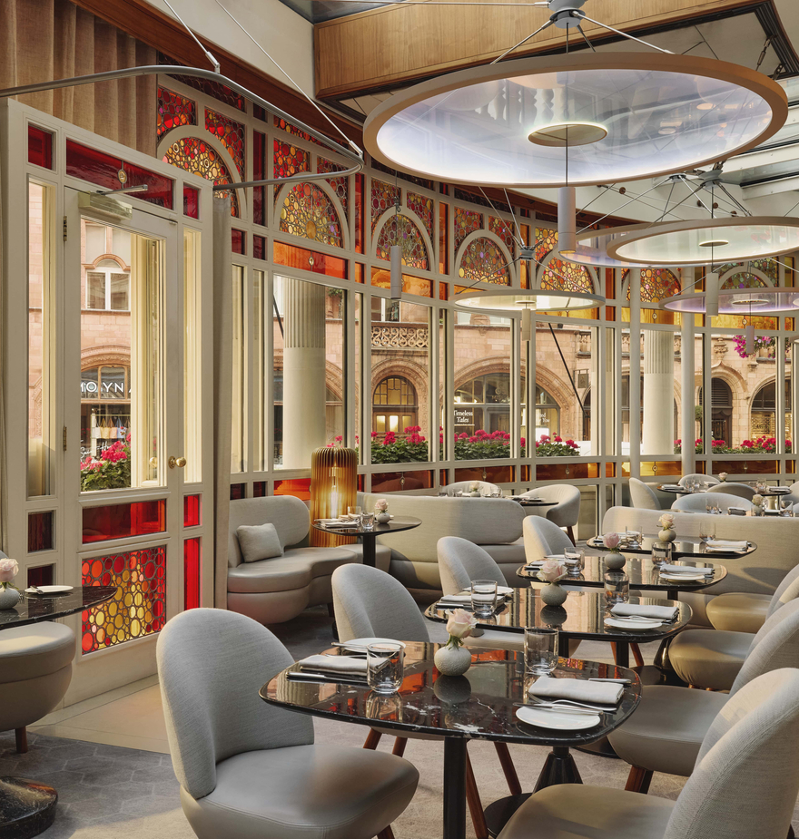 Sunlit restaurant with marble tables, curving banquettes, and stained-glass windows, offering views of Mount Street through red and amber arched panels.