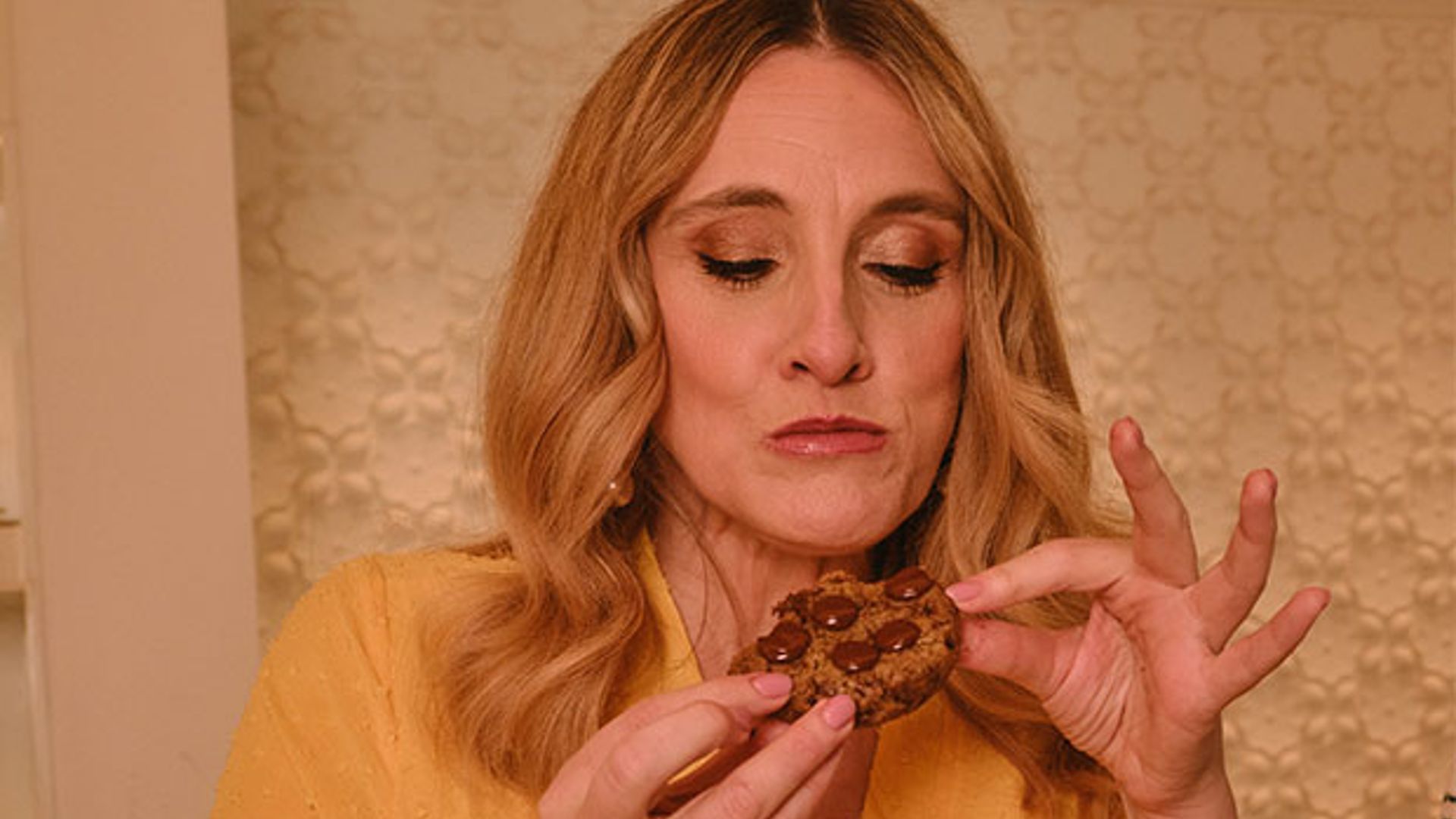 A guest in a vibrant yellow wrap dress savoring a rich chocolate chip cookie, eyes closed in enjoyment, surrounded by an inviting pastry selection.