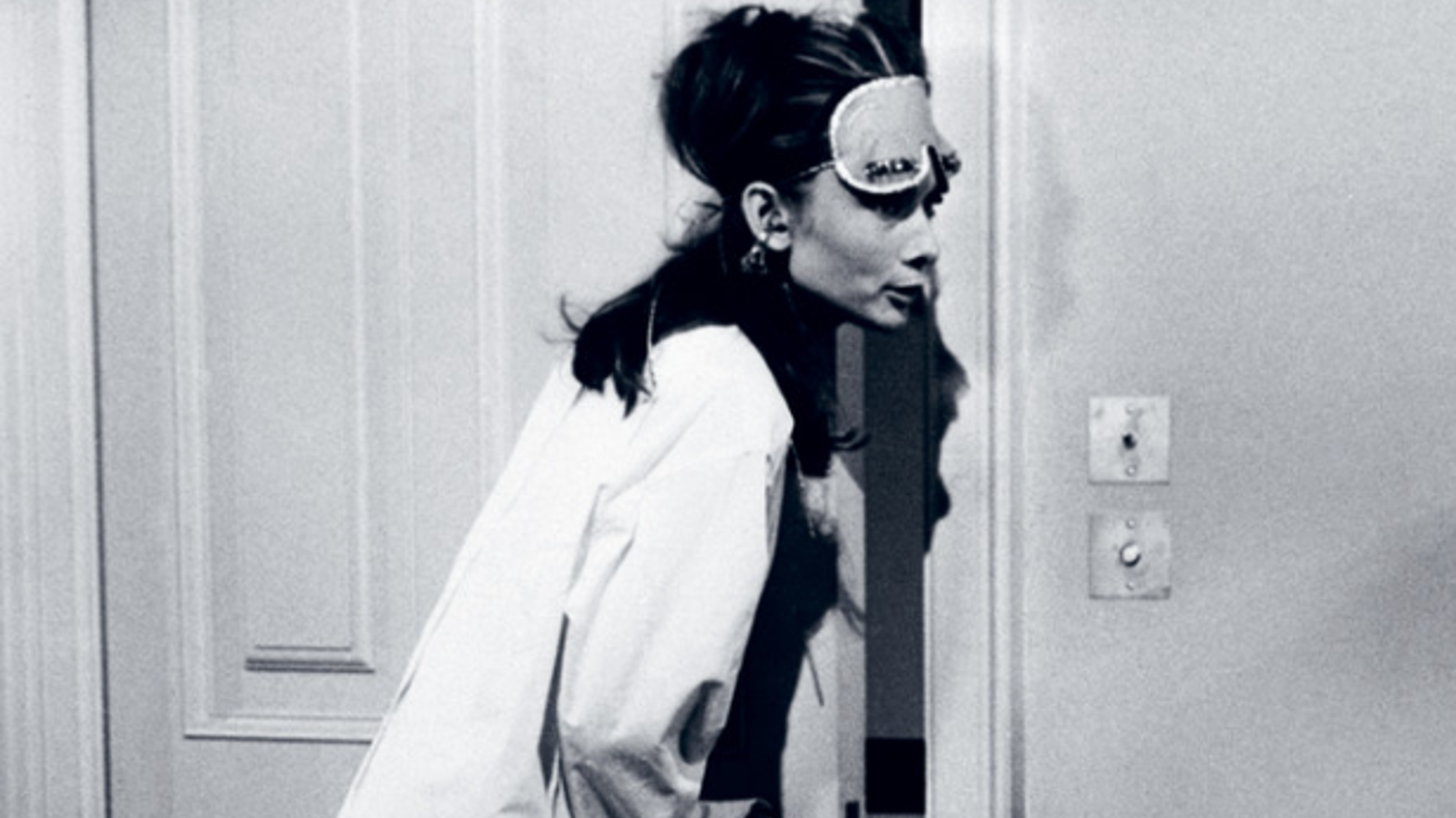 A woman wearing a sleep mask and oversized shirt leans curiously through a partially opened door in a dimly lit room.
