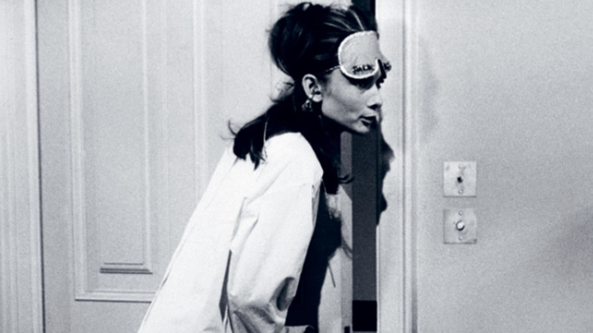 A woman wearing a sleep mask and oversized shirt leans curiously through a partially opened door in a dimly lit room.