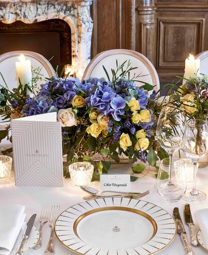 Close-up of a formal table setting with gold-rimmed plates, candles, and purple and yellow floral arrangement at Claridge’s.