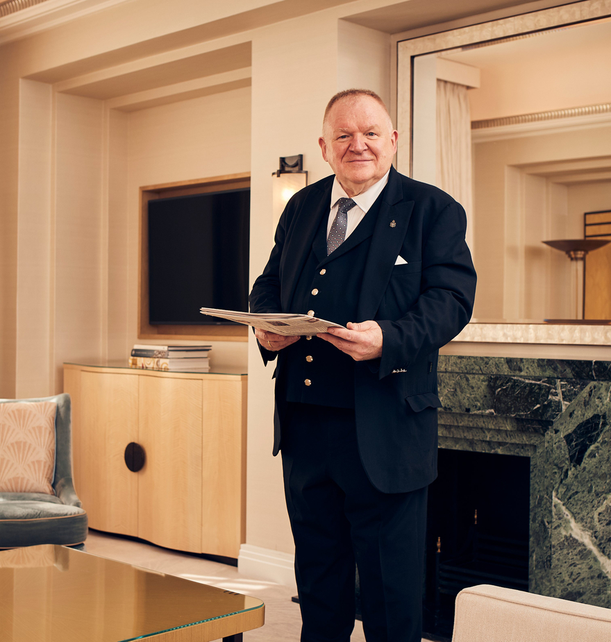 Distinguished butler in a tailored uniform holding a document, standing in an elegant, softly lit luxury suite.