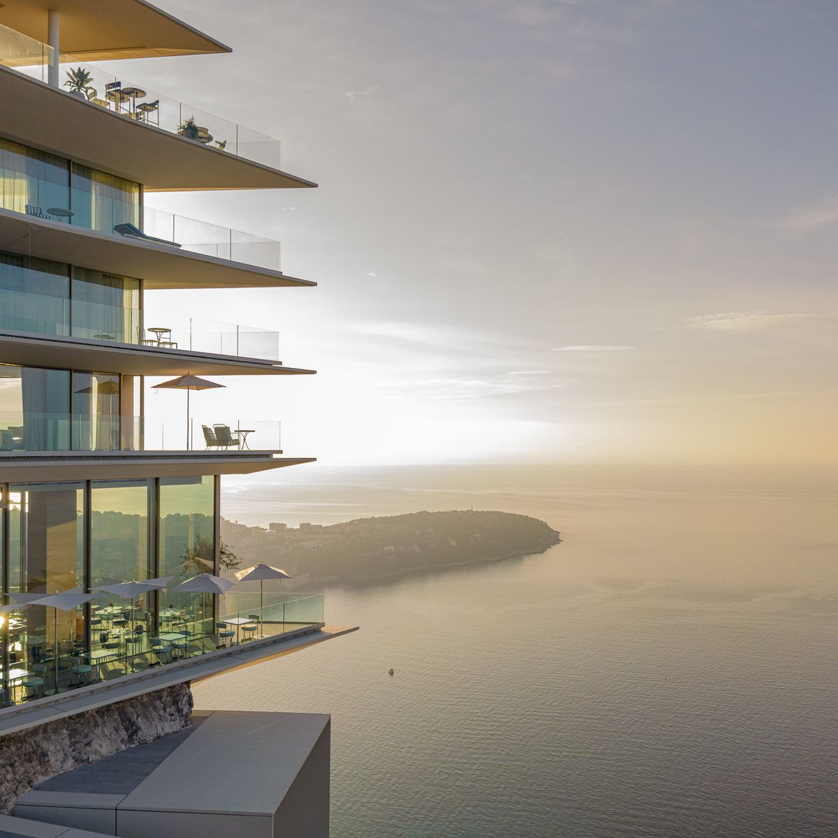 Luxury seafront residence with tiered balconies, glass walls, and sunset reflections over the Mediterranean coastline.