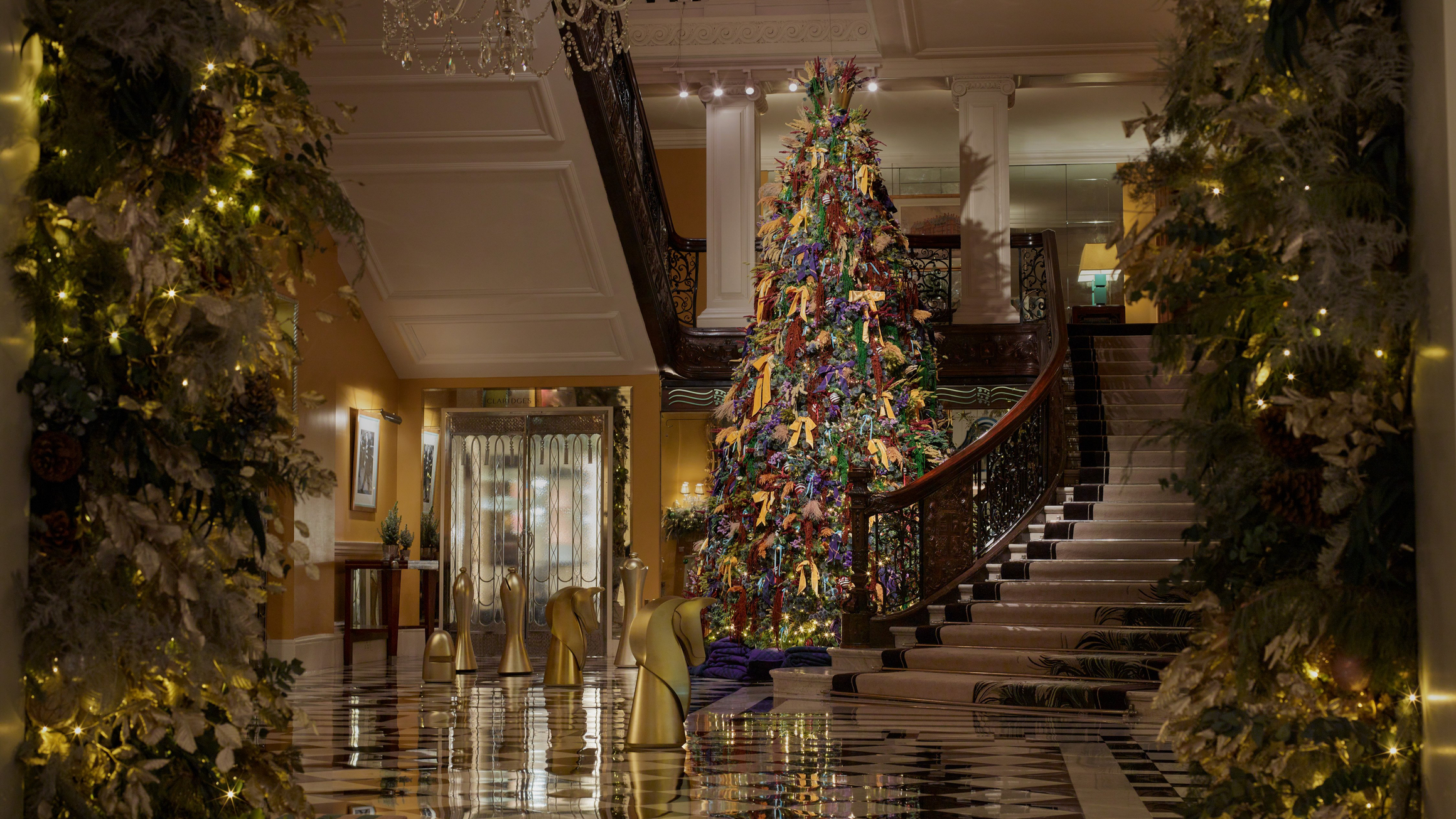 Festive lobby view framed by garlands, showcasing a tall Christmas tree beside a curved staircase and gold chess pieces.