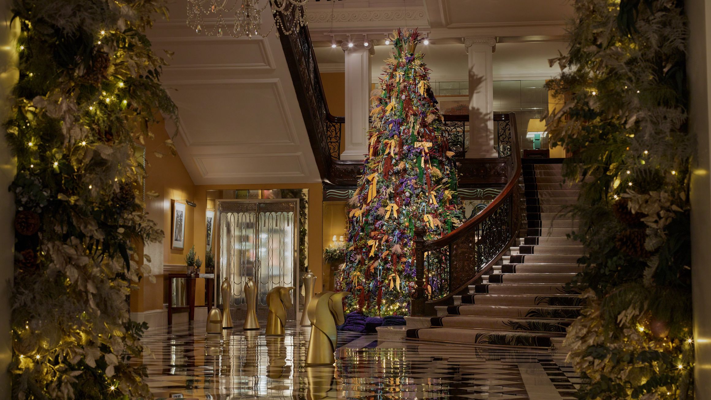 Festive lobby view framed by garlands, showcasing a tall Christmas tree beside a curved staircase and gold chess pieces.