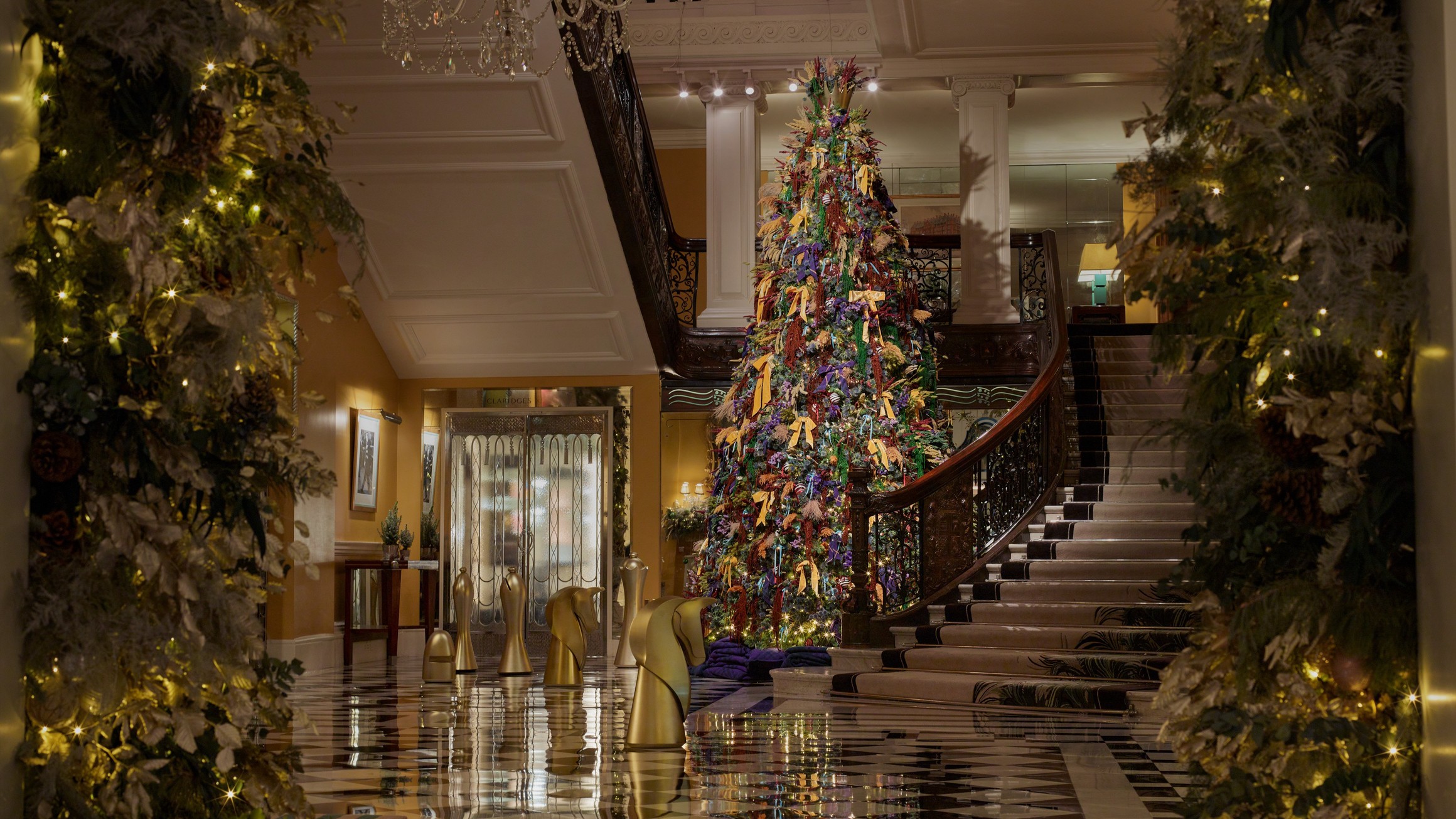 Festive lobby view framed by garlands, showcasing a tall Christmas tree beside a curved staircase and gold chess pieces.
