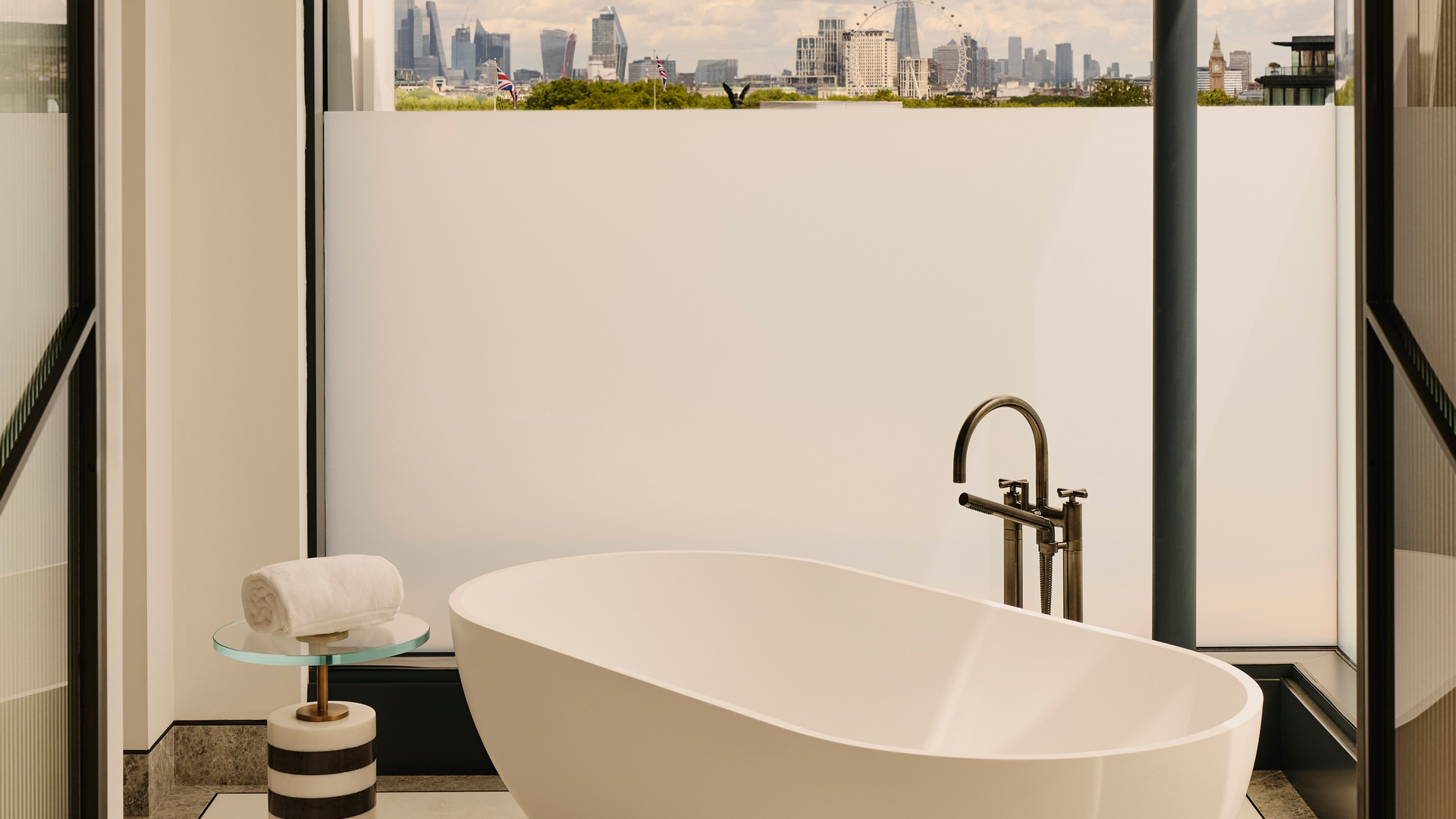 Freestanding white bathtub in marble bathroom, facing skyline view of London through floor-to-ceiling glass, including The Shard and London Eye.