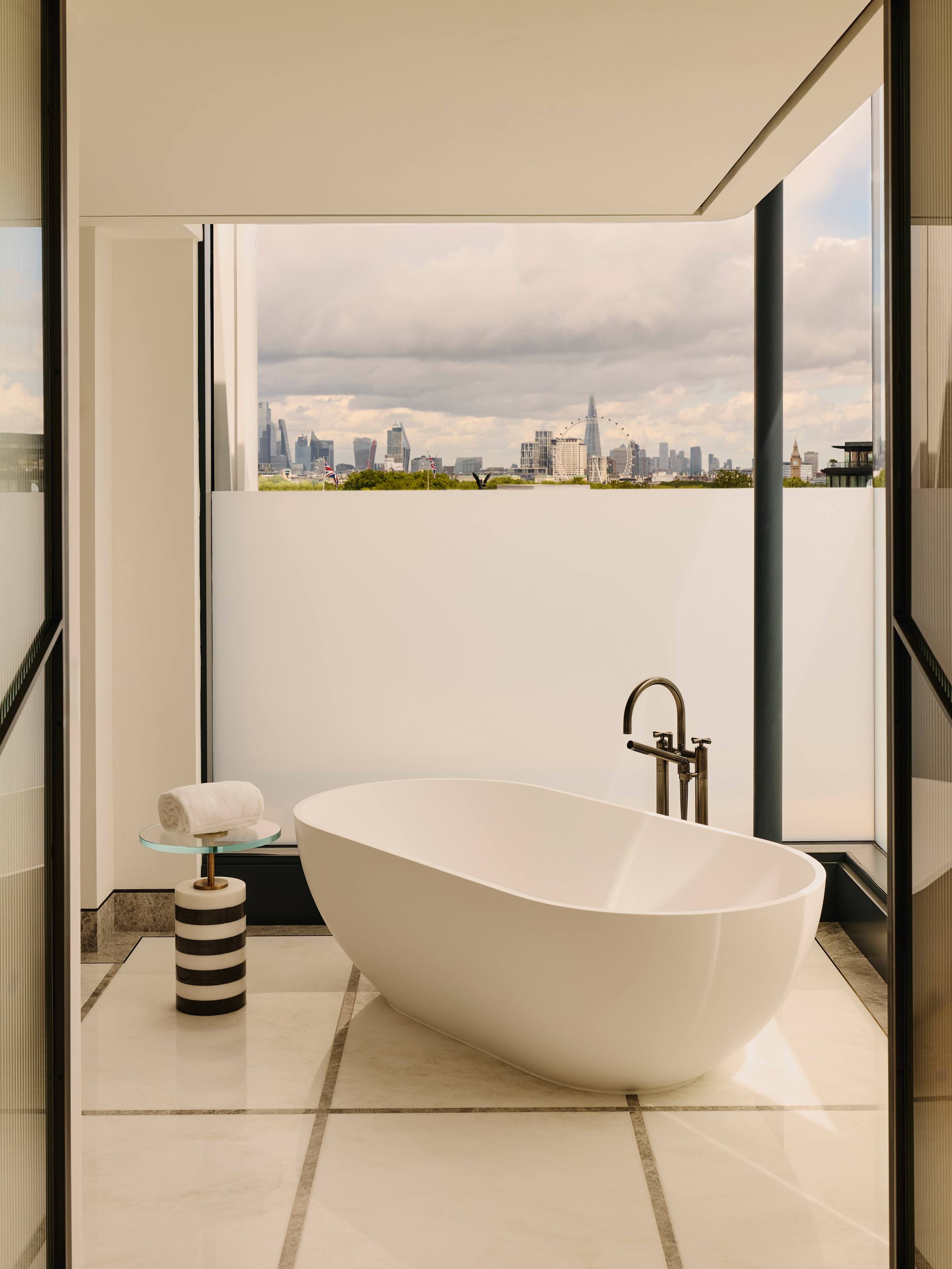Freestanding white bathtub in marble bathroom, facing skyline view of London through floor-to-ceiling glass, including The Shard and London Eye.