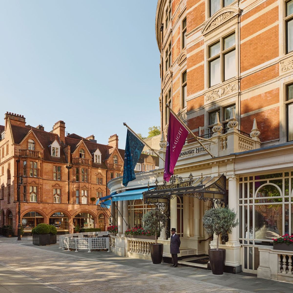 The elegant exterior of The Connaught Hotel, featuring red-brick architecture, flags, and a doorman under a decorative entrance.