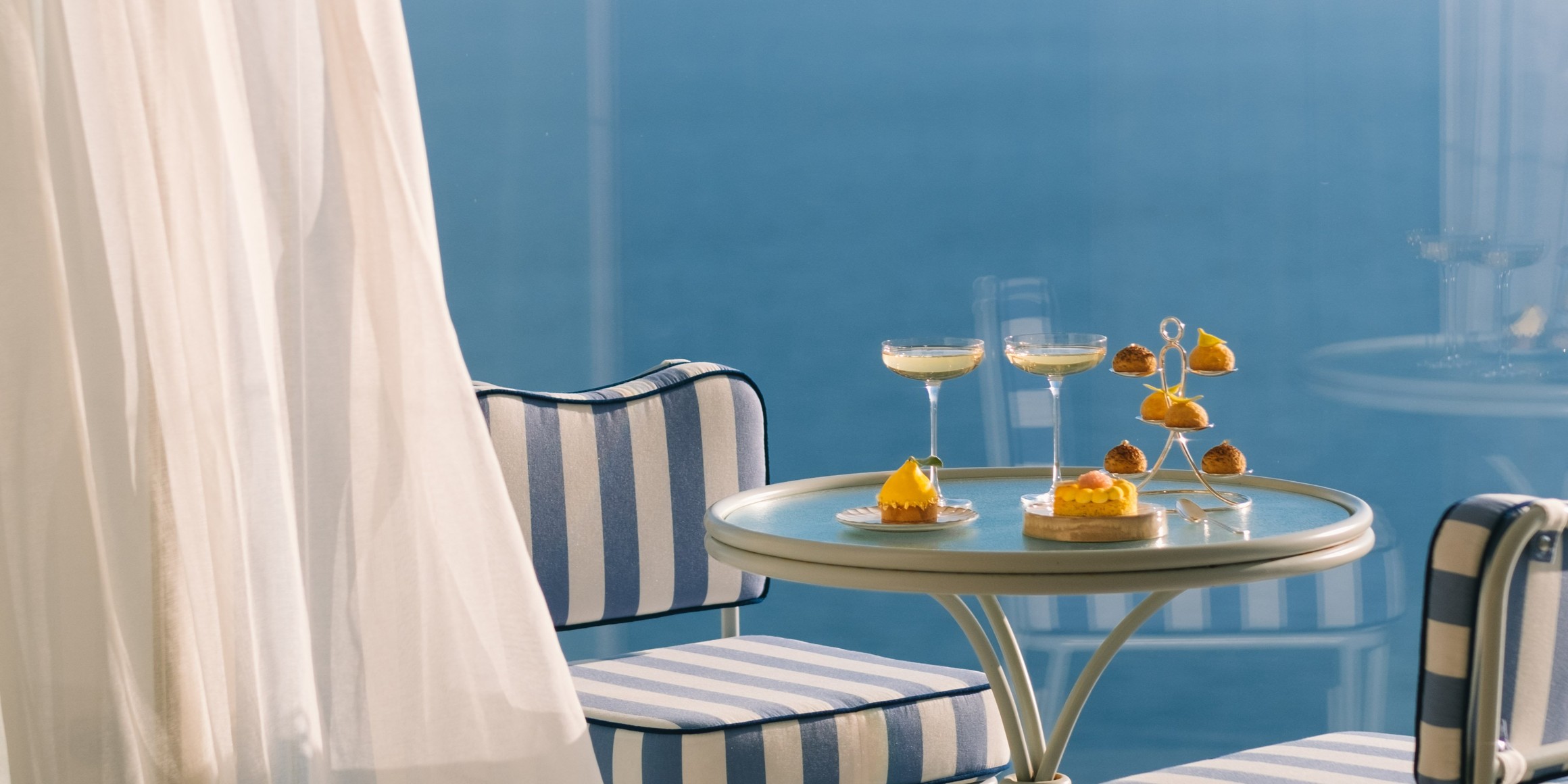 A serene seaside balcony with striped chairs, a small table, and elegant desserts overlooking a tranquil blue ocean.