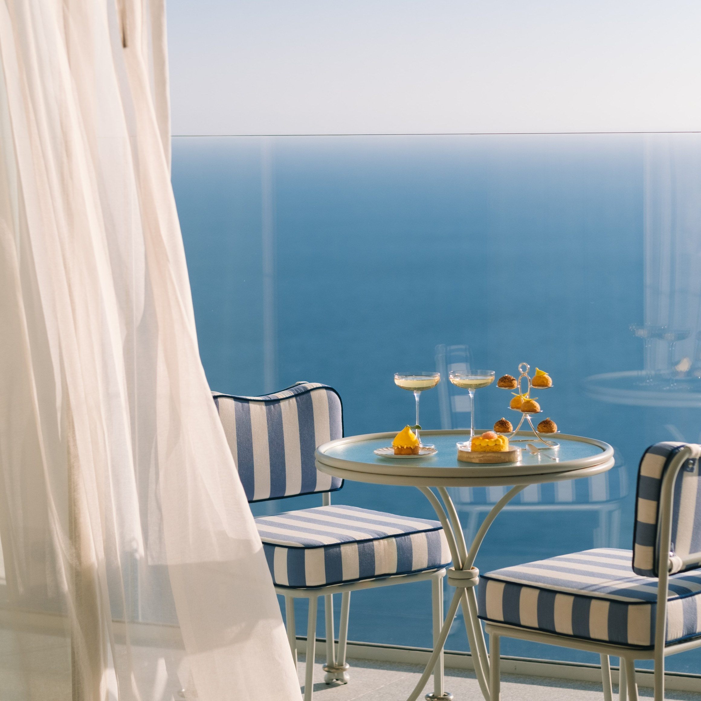 A serene seaside balcony with striped chairs, a small table, and elegant desserts overlooking a tranquil blue ocean.