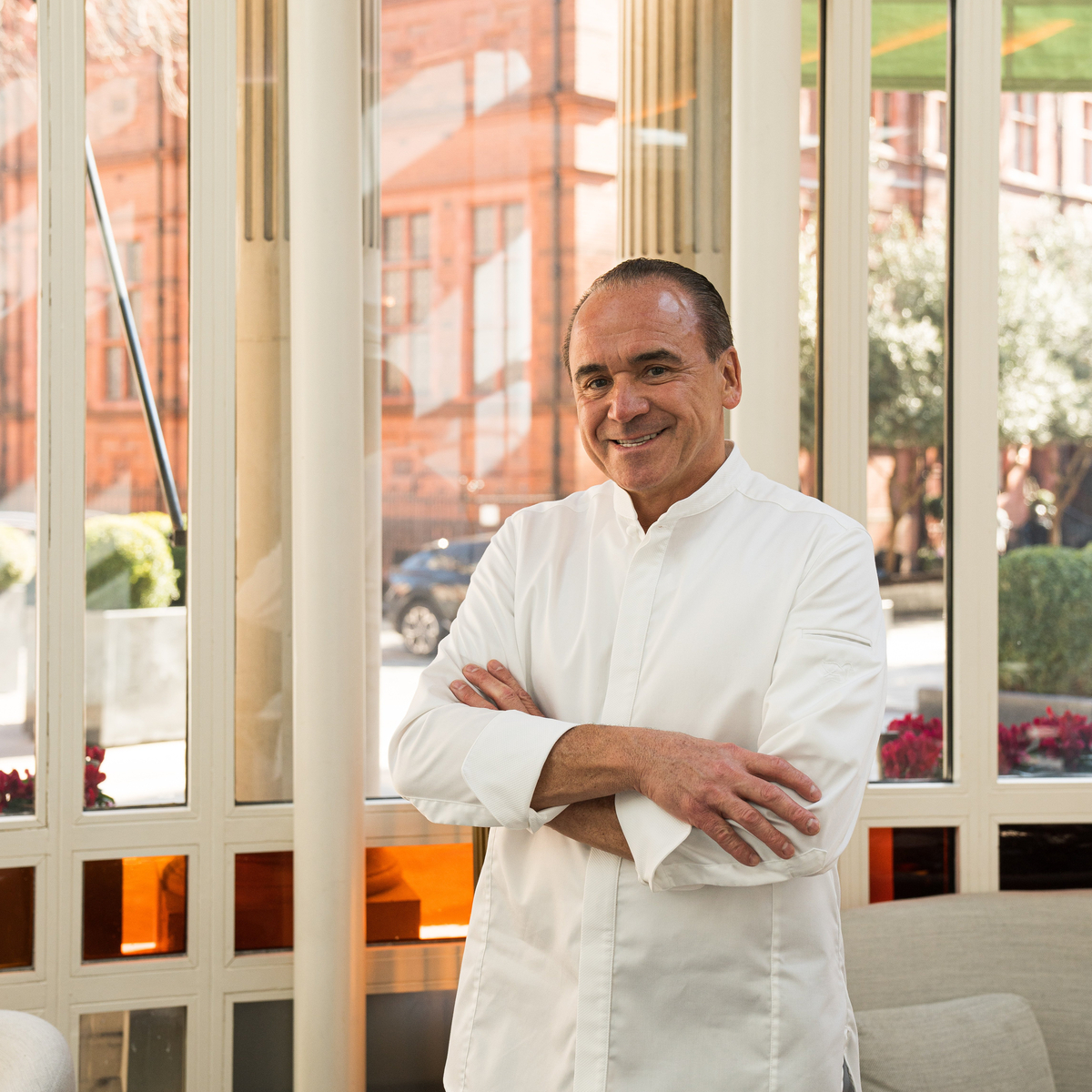 Chef in a white jacket standing with arms crossed, smiling in front of stained-glass windows with orange accents and a city street view.