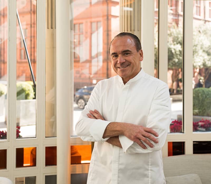 Chef in a white jacket standing with arms crossed, smiling in front of stained-glass windows with orange accents and a city street view.