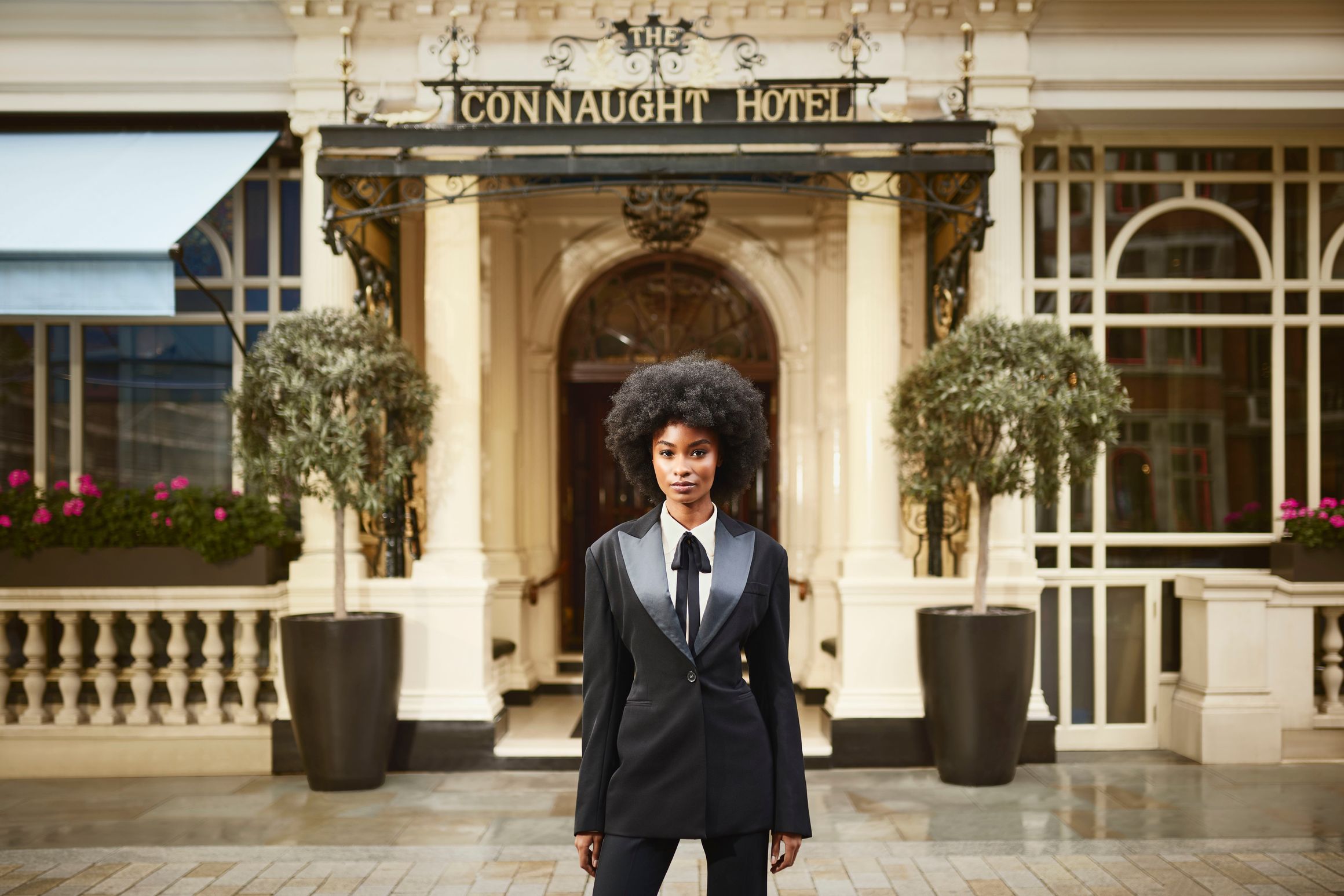 A confident individual in a tailored black suit with satin lapels stands gracefully outside The Connaught Hotel's grand entrance,