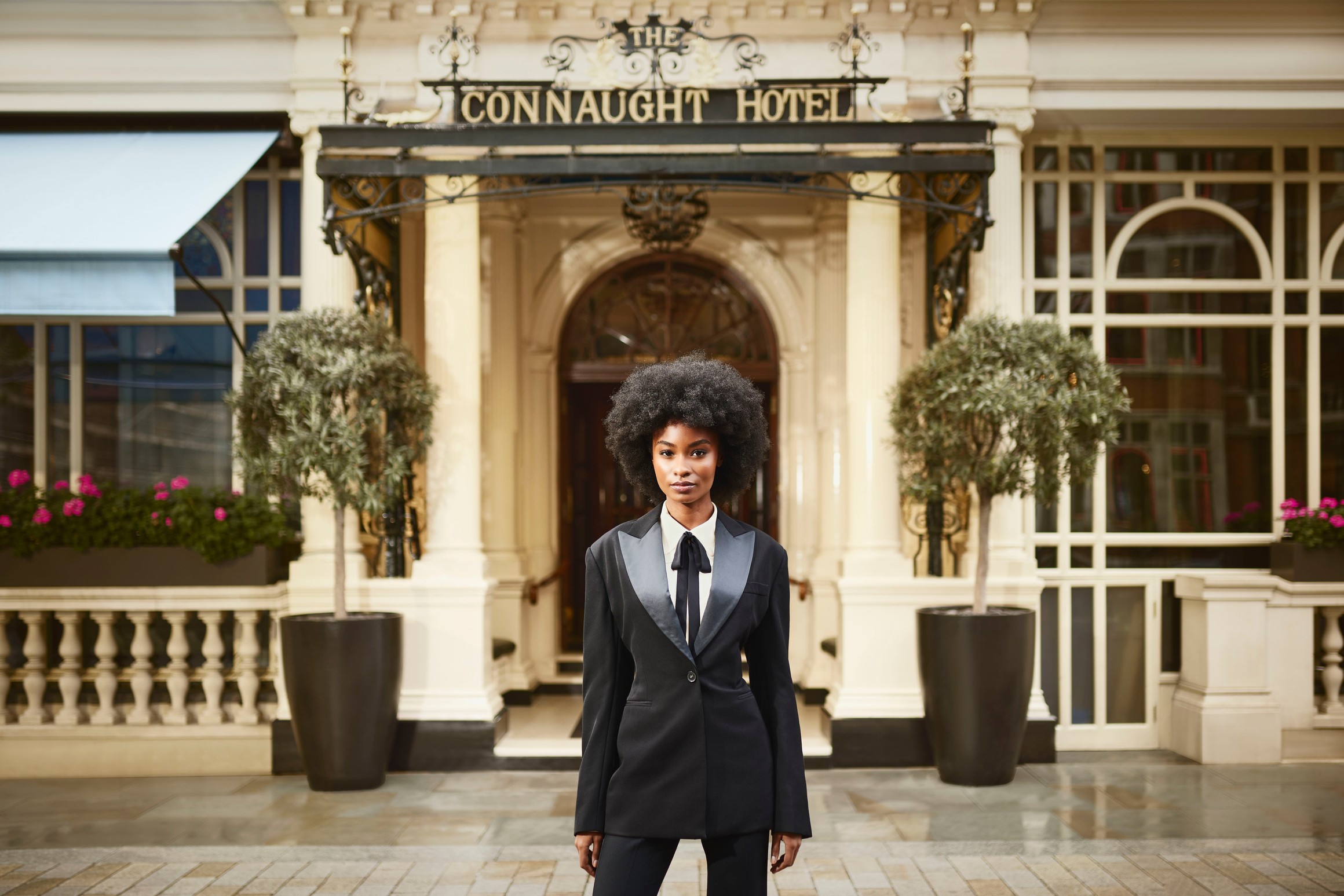 A confident individual in a tailored black suit with satin lapels stands gracefully outside The Connaught Hotel's grand entrance,