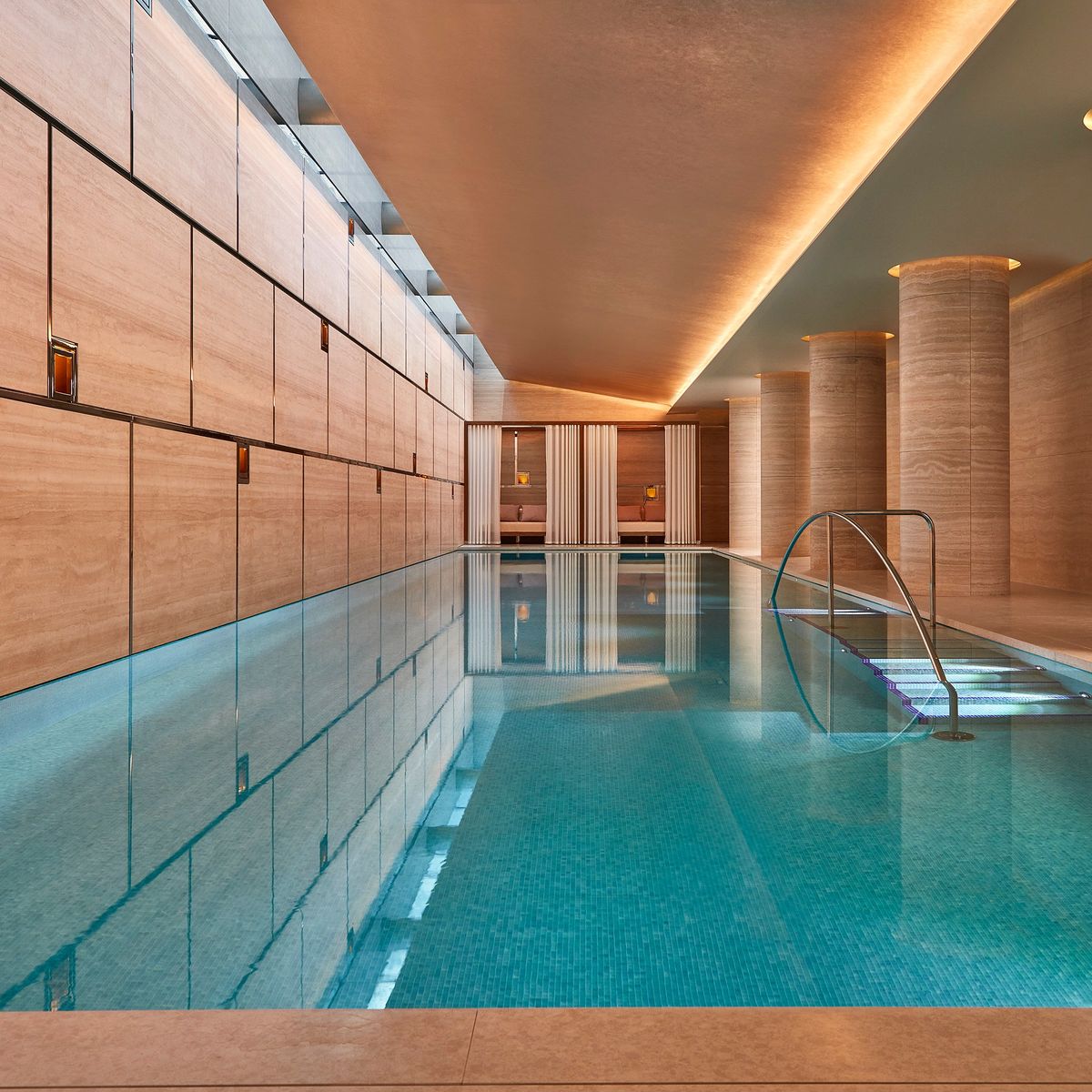 A serene indoor swimming pool with warm-toned stone walls, soft lighting, and elegant columned architecture creating a tranquil retreat.