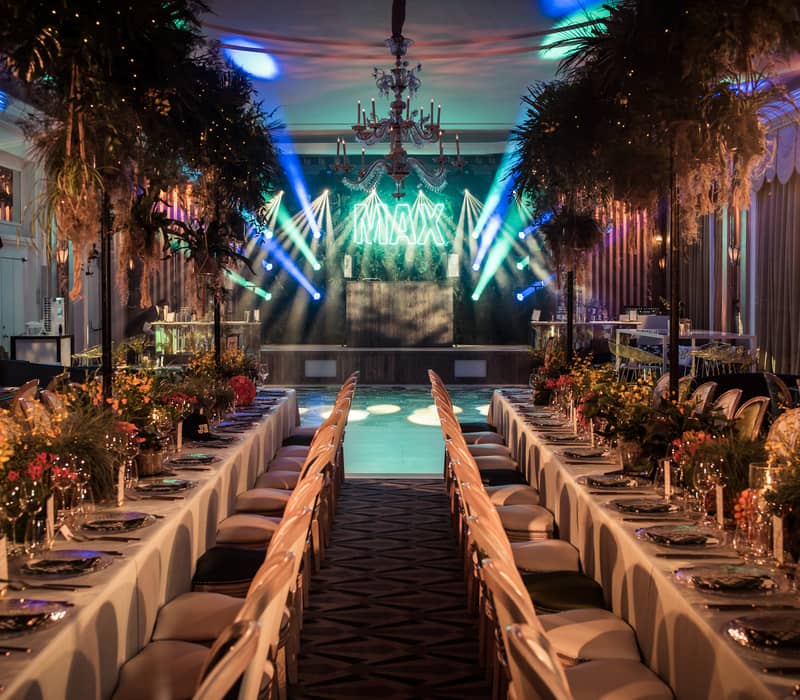 Lavish event space with long banquet tables, lush foliage, and vibrant stage lighting for a glamorous celebration.