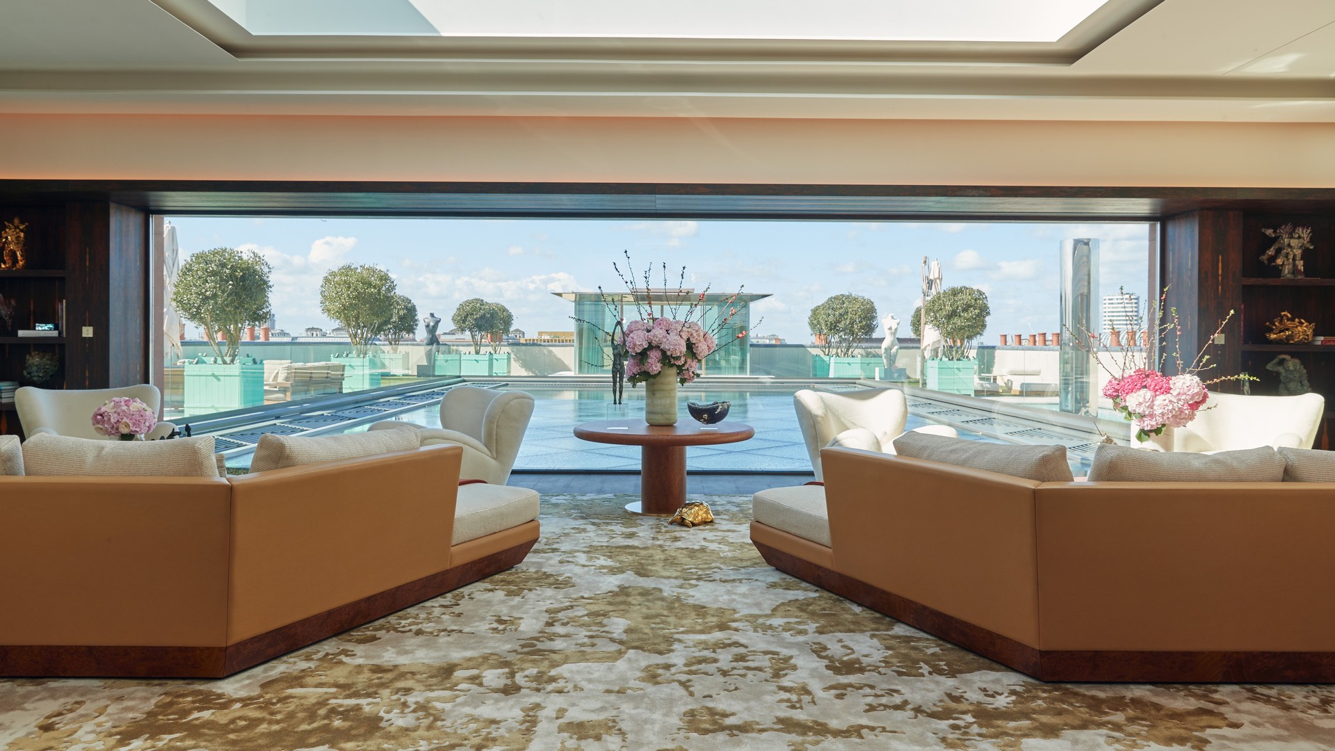 A luxurious sitting room features plush seating and elegant floral arrangements, offering a stunning view of a rooftop pool and manicured greenery under a bright sky.
