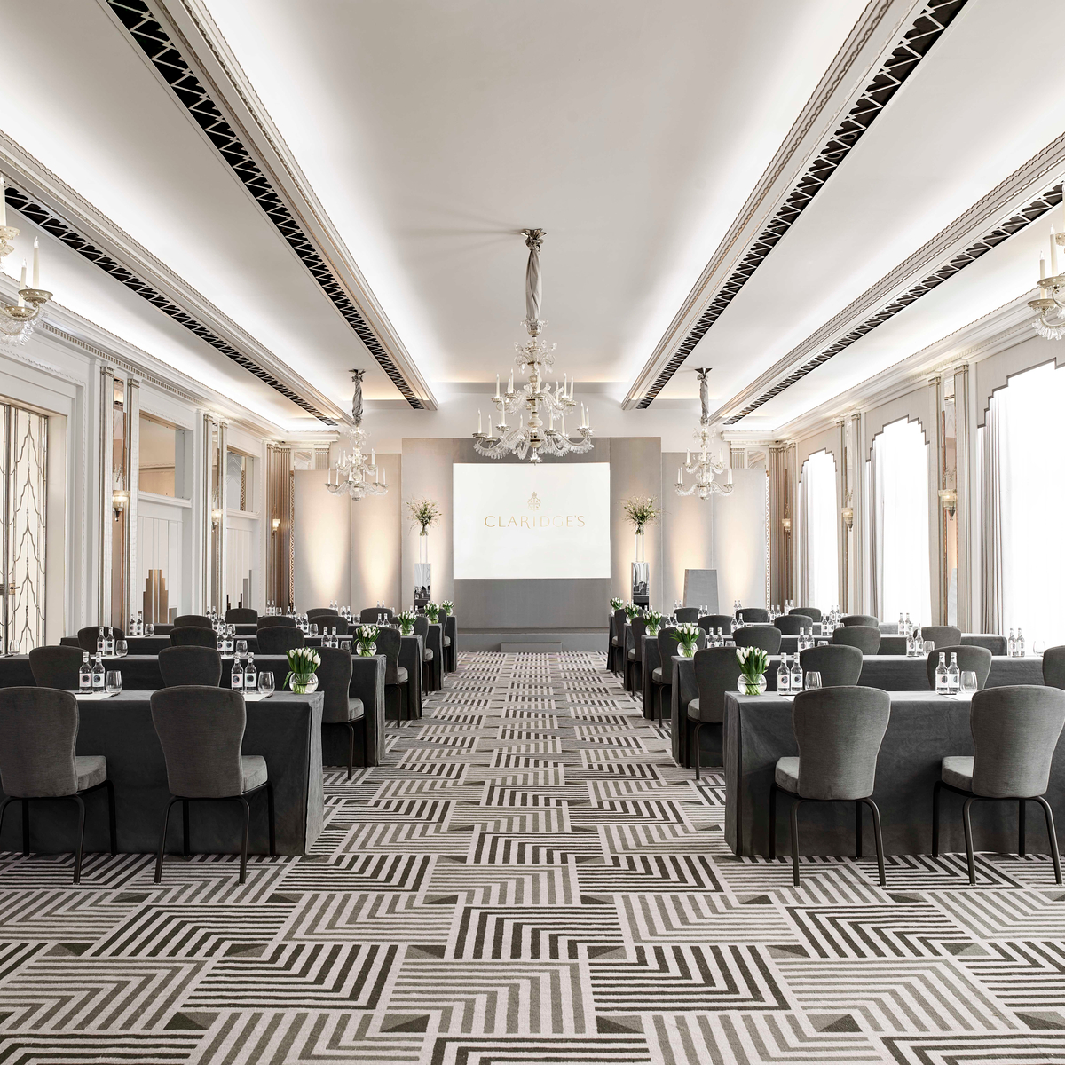 Luxurious conference room with elegant chandeliers, natural light, geometric carpet design, and neatly arranged tables and chairs.