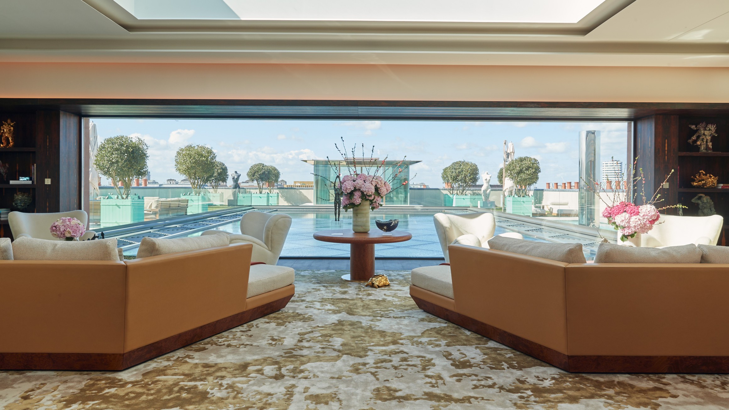 A luxurious sitting room features plush seating and elegant floral arrangements, offering a stunning view of a rooftop pool and manicured greenery under a bright sky.