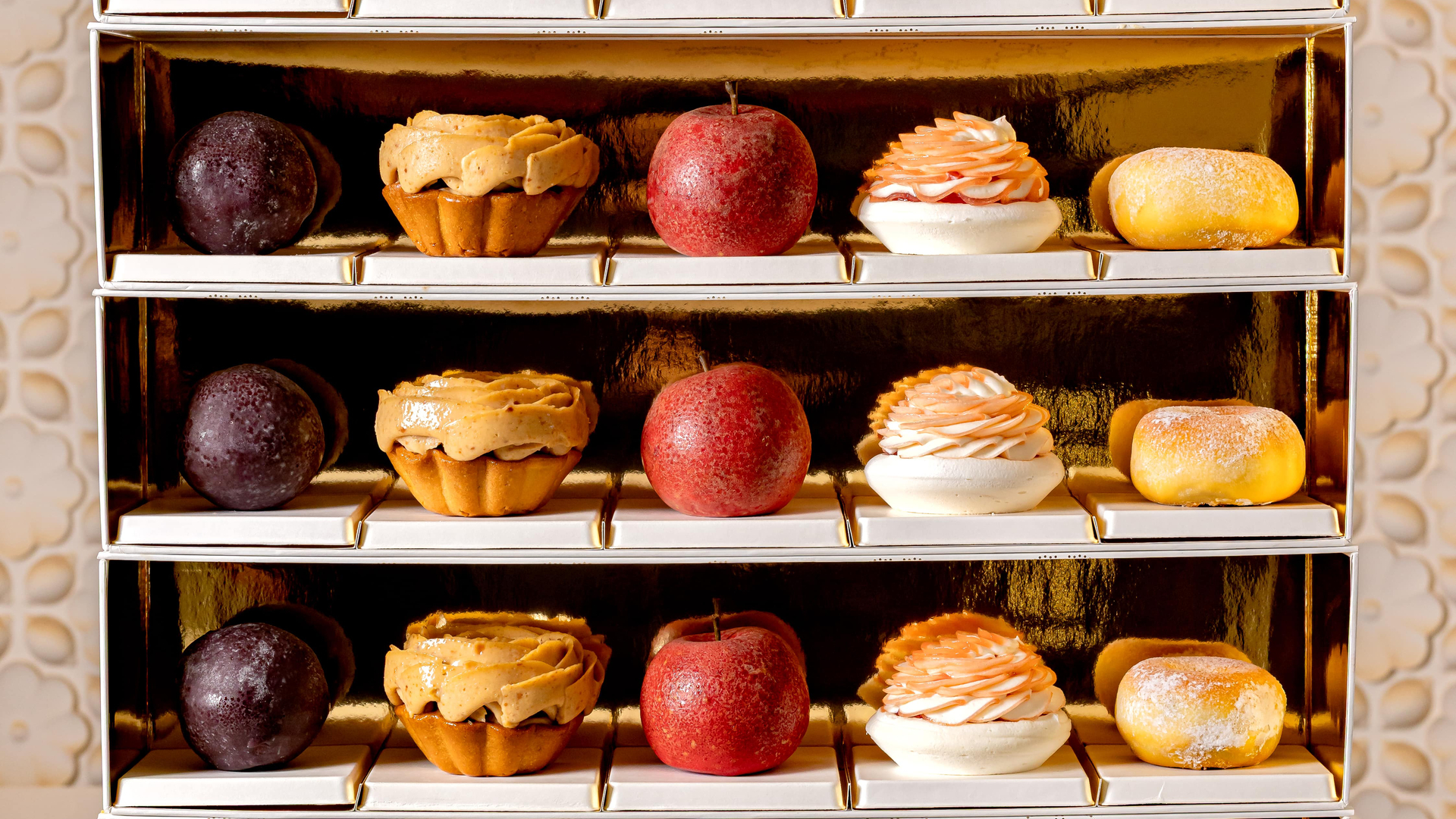 Elegant pastry presentation box with four gold-lined shelves, each featuring a symmetrical arrangement of fruit-inspired desserts, tartlets, and meringue creations, set against a softly patterned backdrop.