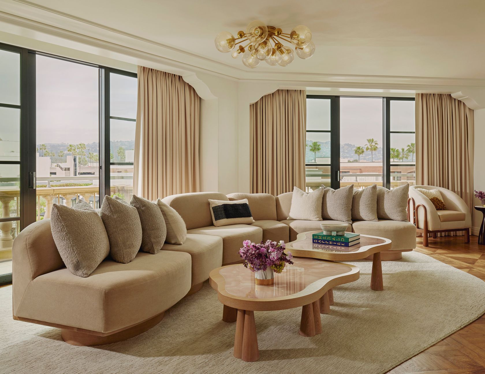 Elegant living room with curved beige sofa, sculptural tables, and large windows overlooking palm trees and hills beyond.
