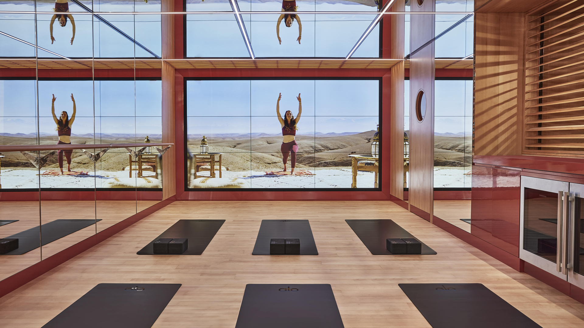 Modern mirrored yoga studio with virtual instructor on desert landscape screen and mats arranged on wooden floor.