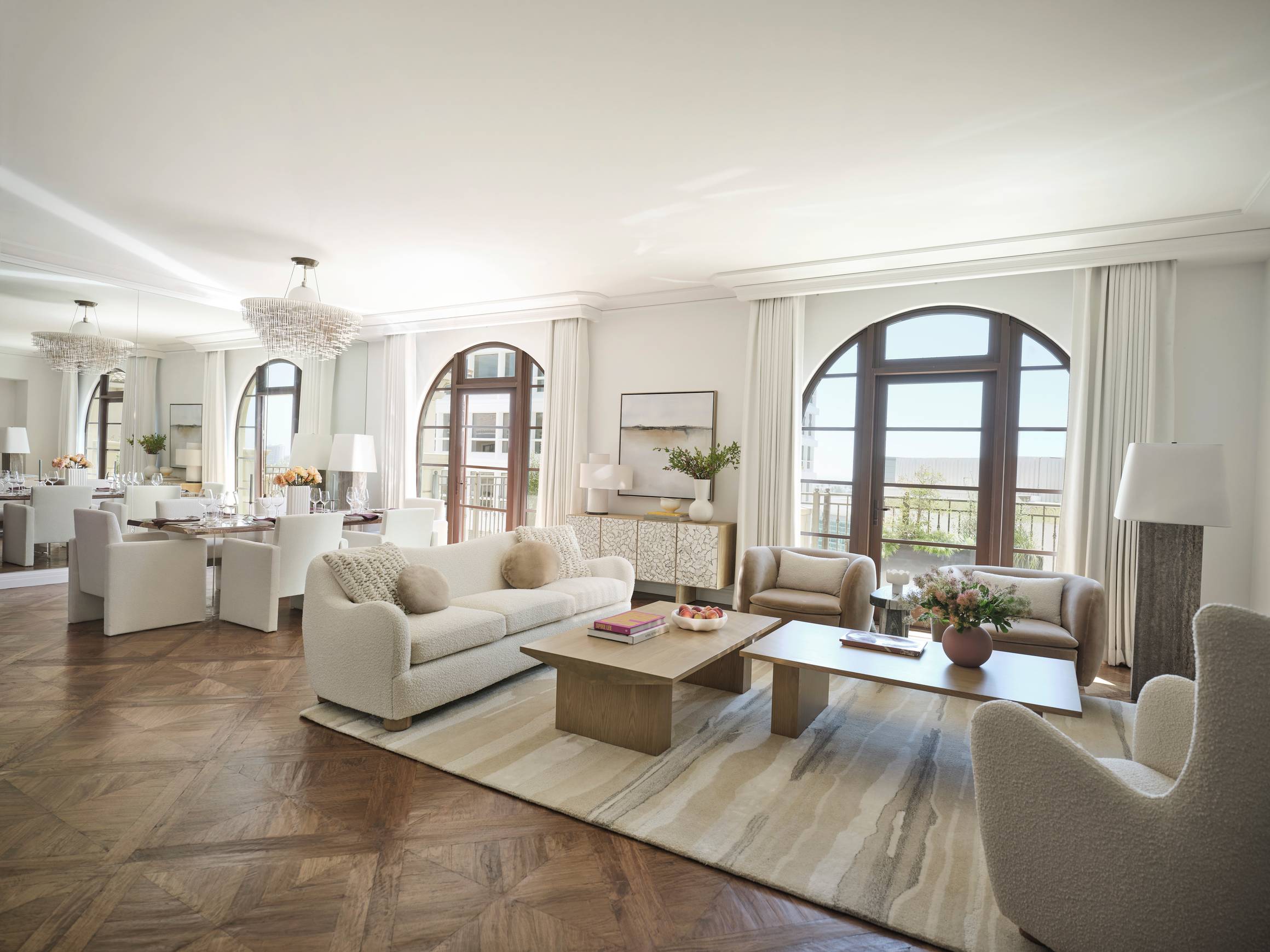 A sophisticated open-plan living and dining room with arched windows, crystal chandeliers, and plush furnishings in a neutral palette.