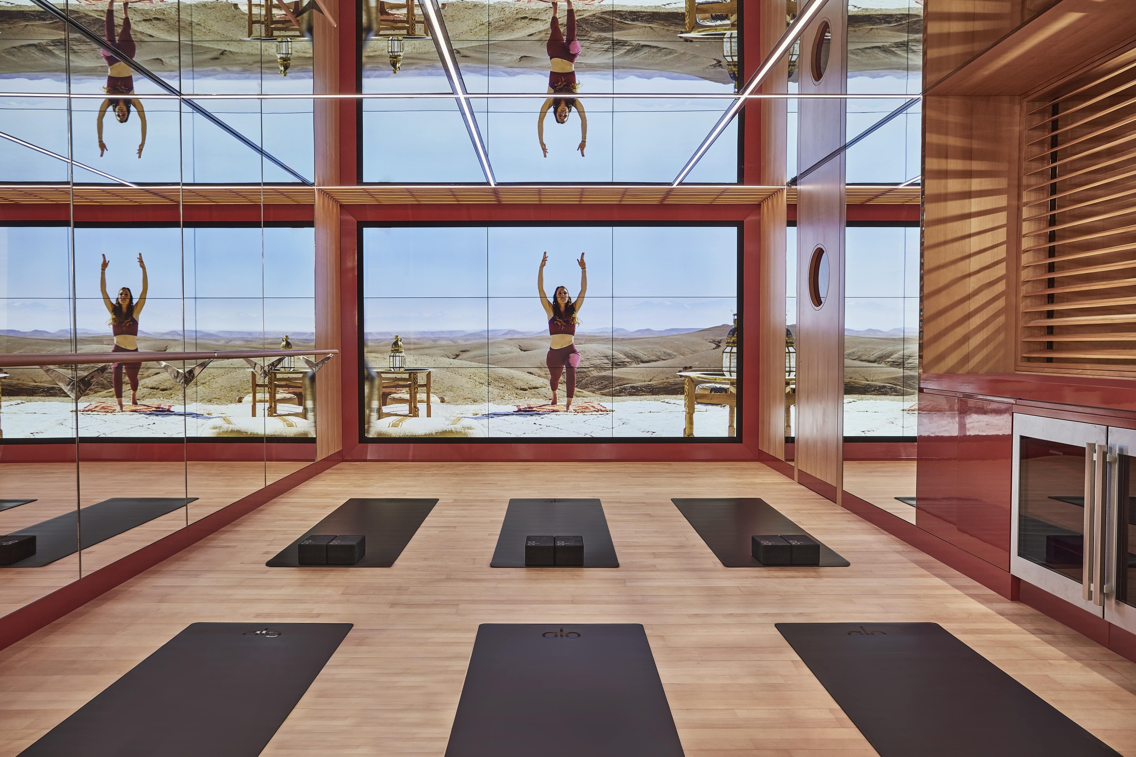 Modern mirrored yoga studio with virtual instructor on desert landscape screen and mats arranged on wooden floor.