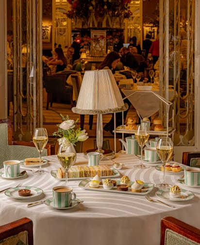 Table set for afternoon tea with striped china, patisserie, and champagne, in an elegant, mirrored salon with floral centerpiece and warm lighting.