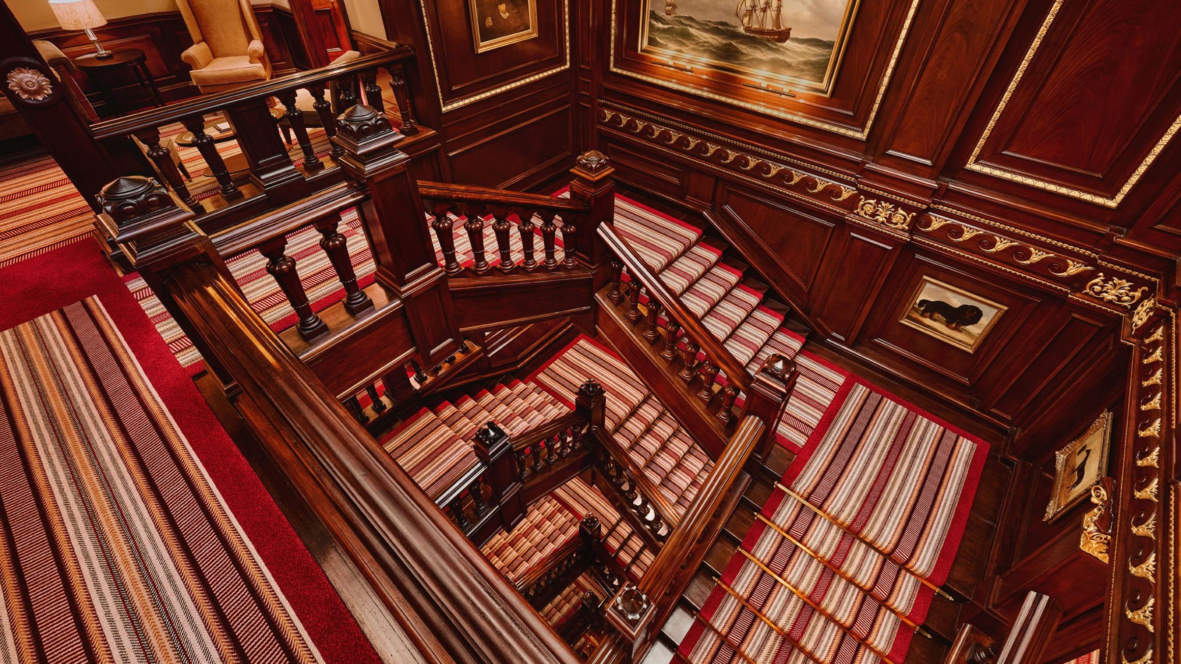 Ornate multi-level wooden staircase with striped red and beige carpet runners, viewed from above.