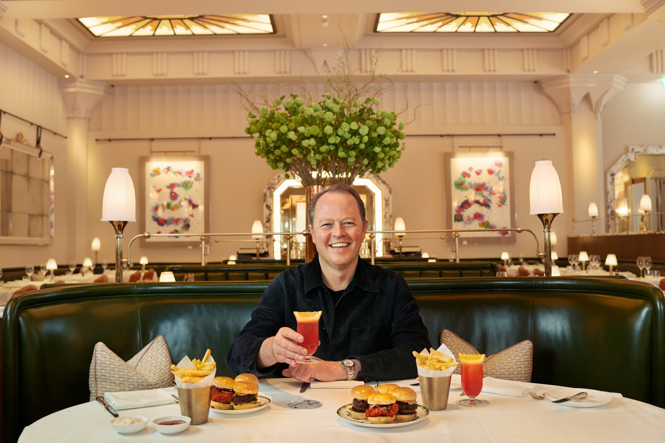A smiling man in Claridge’s restaurant raises a cocktail, with mini burgers and fries on the table.