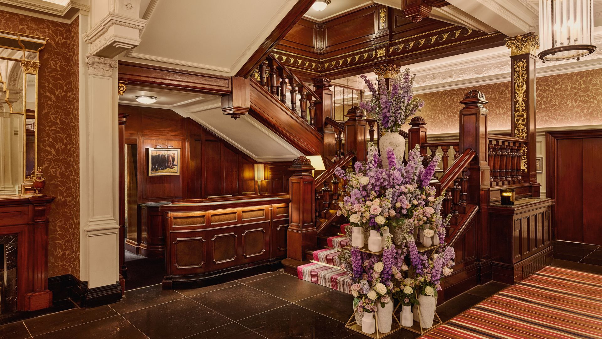 Ornate hotel lobby with dark wood paneling, grand staircase, and a large floral arrangement featuring purple and cream flowers.