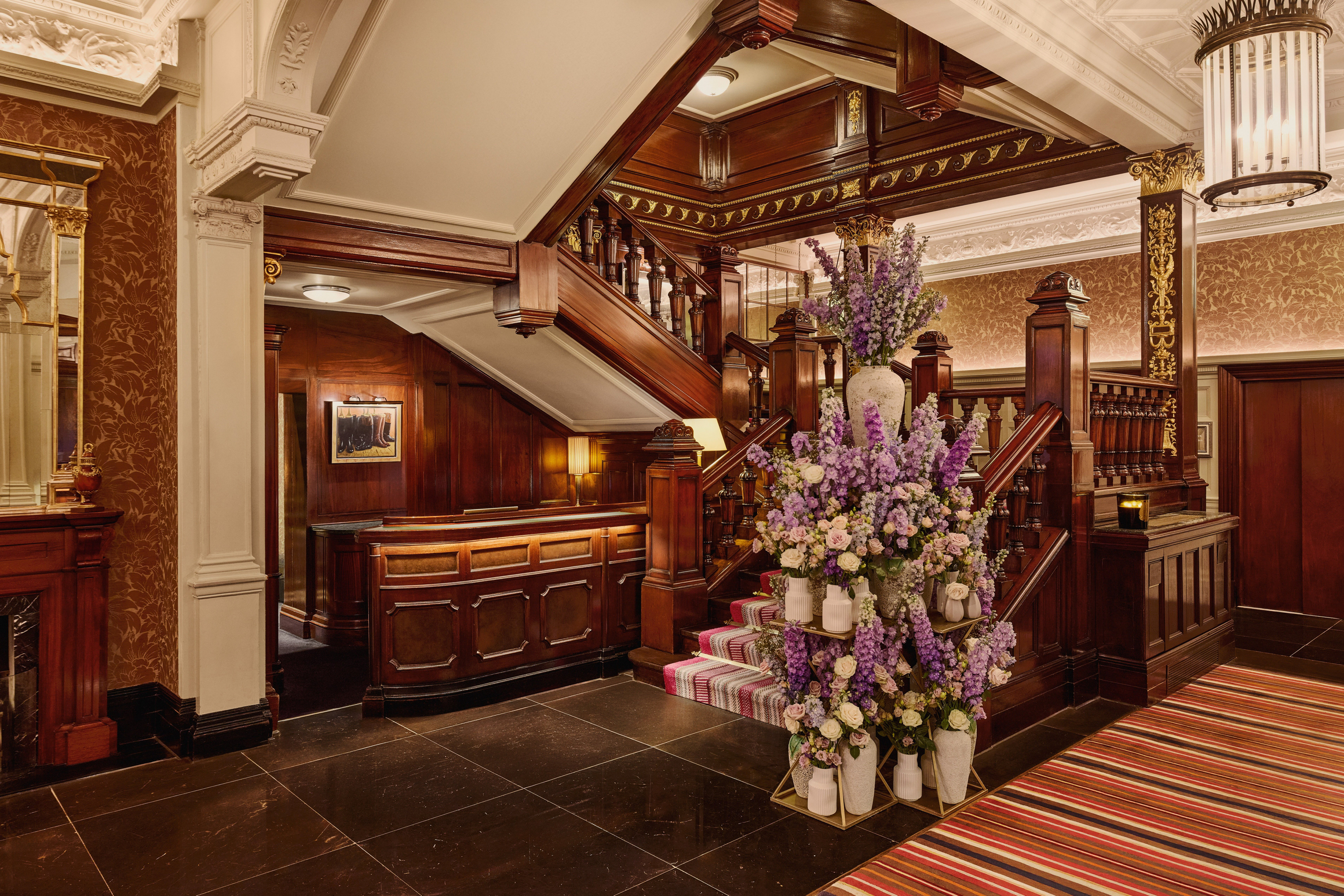 Ornate hotel lobby with dark wood paneling, grand staircase, and a large floral arrangement featuring purple and cream flowers.