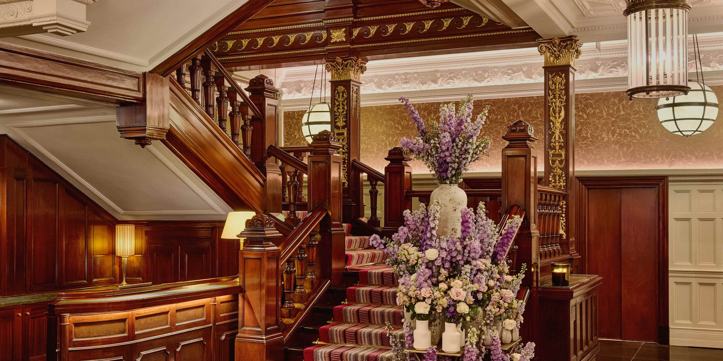Grand staircase with dark wood paneling, ornate ceiling, striped carpet, and a tall floral arrangement featuring purple and white flowers.
