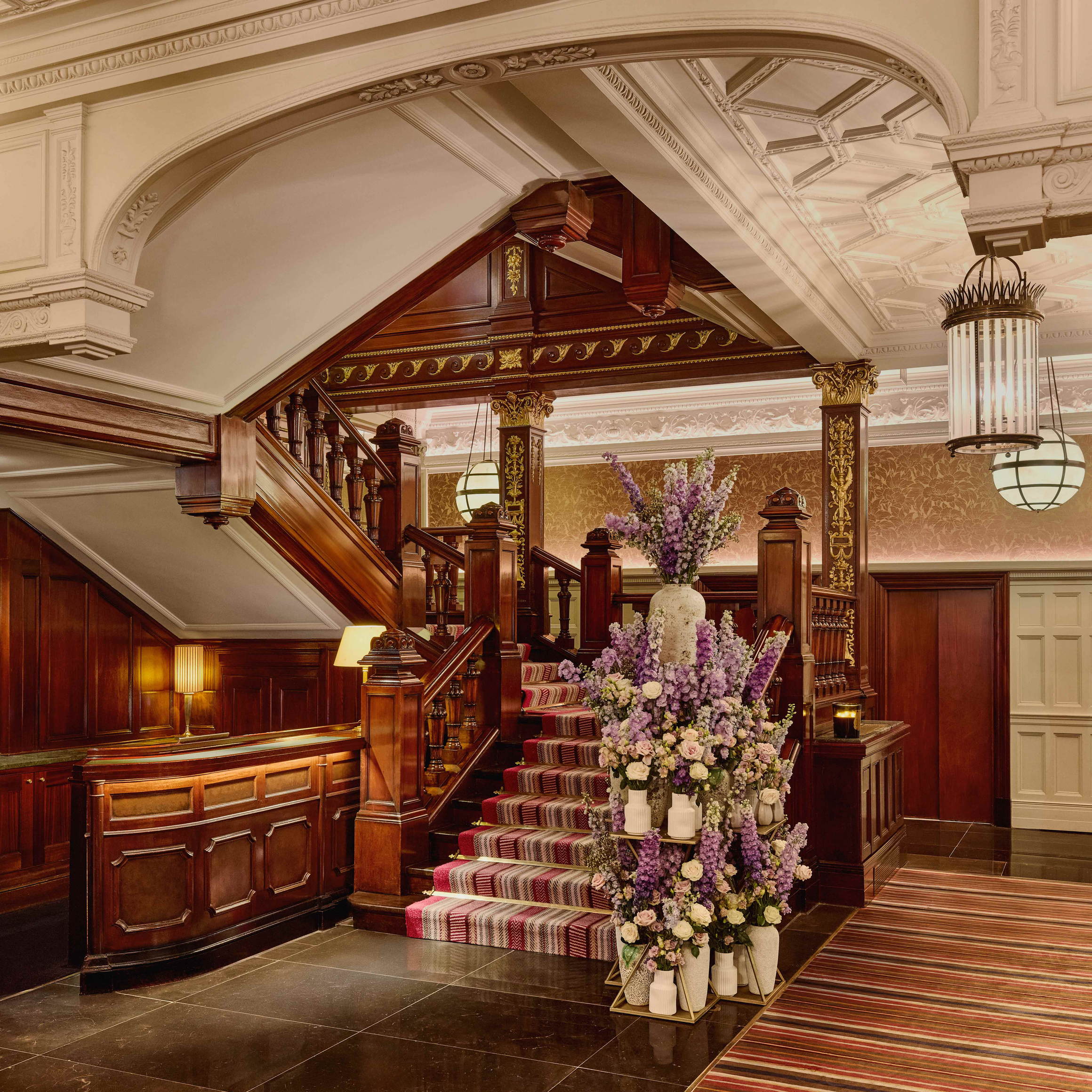 Grand staircase with dark wood paneling, ornate ceiling, striped carpet, and a tall floral arrangement featuring purple and white flowers.