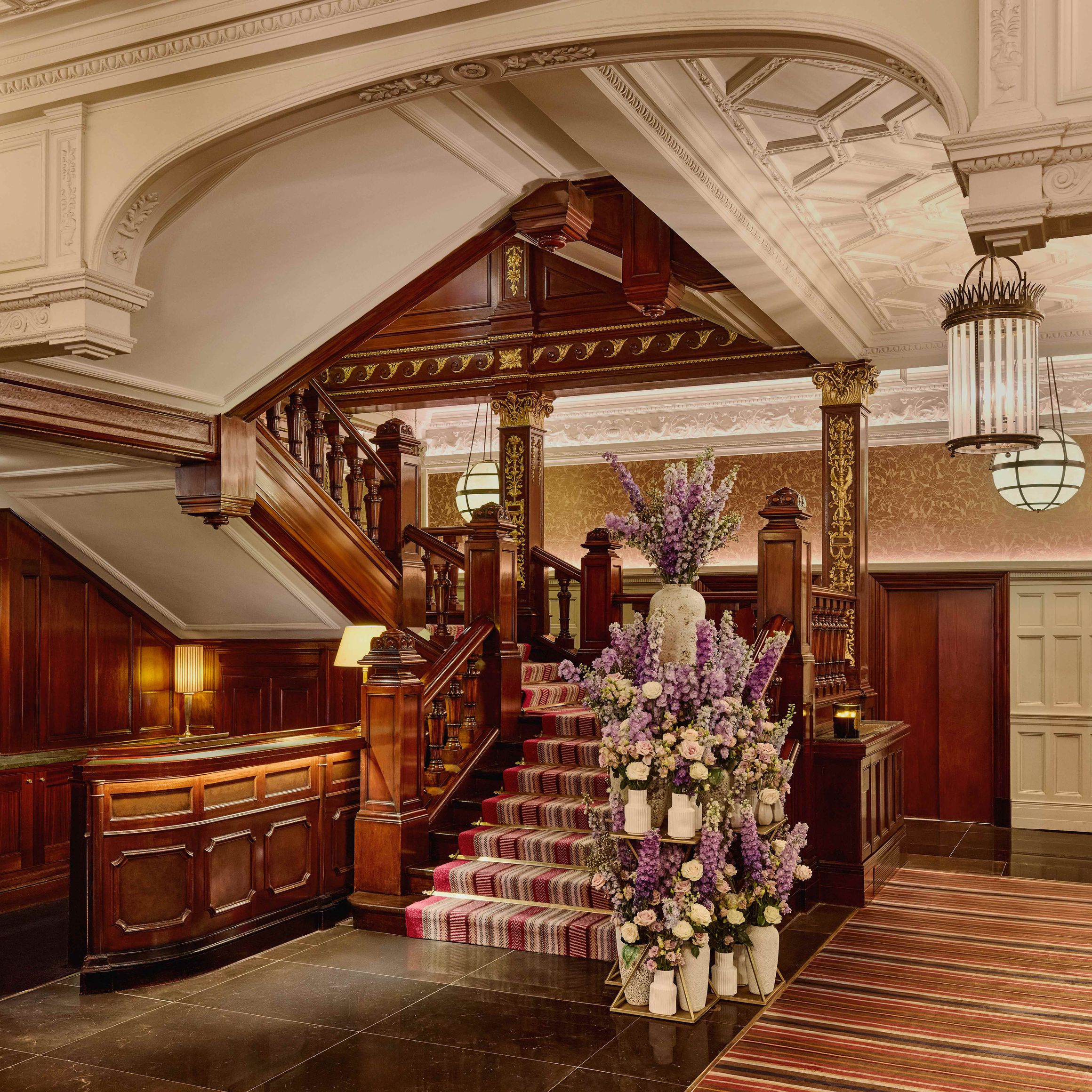 Grand staircase with dark wood paneling, ornate ceiling, striped carpet, and a tall floral arrangement featuring purple and white flowers.