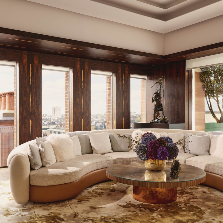 Curved cream sofa in Claridge’s Penthouse surrounds a round table with hydrangeas, set against dark wood panels and city views.