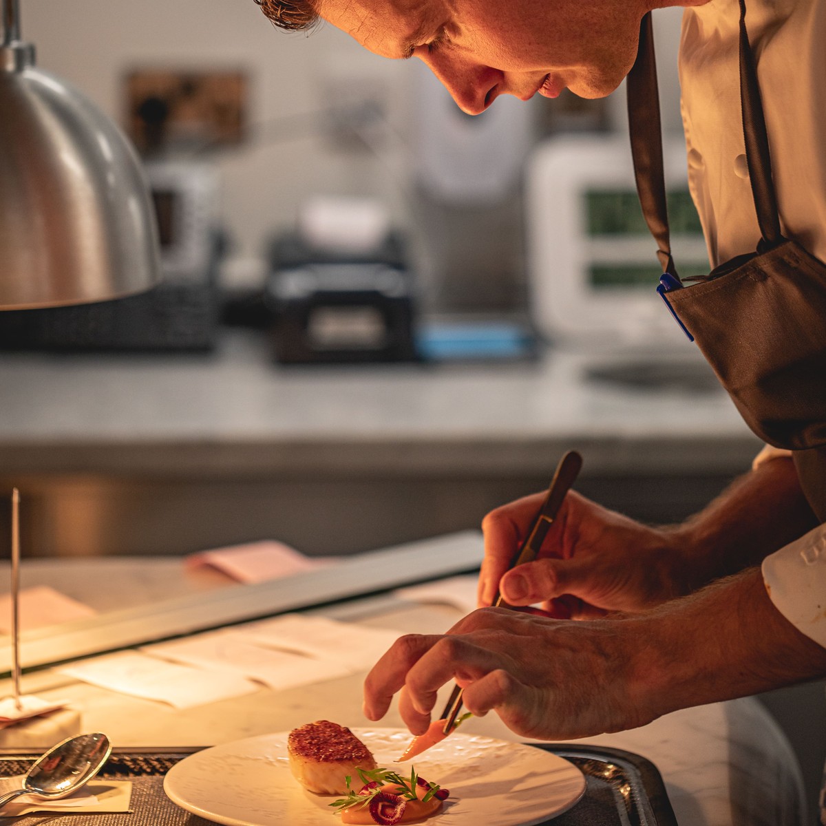 A chef meticulously plates a dish under warm lighting, showcasing precision and artistry in a professional kitchen setting.