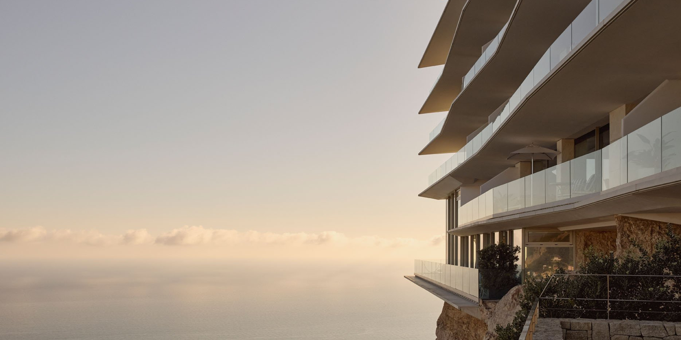 Modern cliffside hotel with cascading glass balconies overlooking a calm, sunlit sea, set against a pastel sky at golden hour.