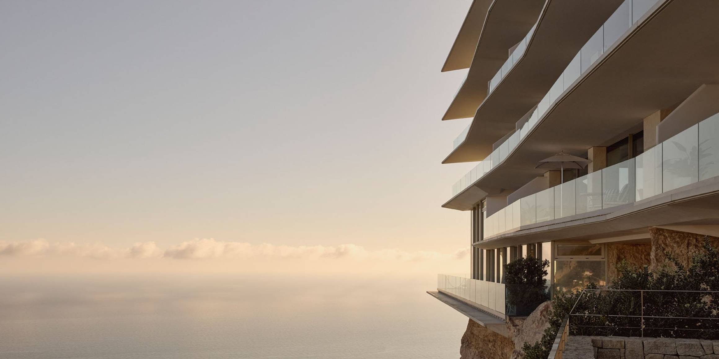 Modern cliffside hotel with cascading glass balconies overlooking a calm, sunlit sea, set against a pastel sky at golden hour.