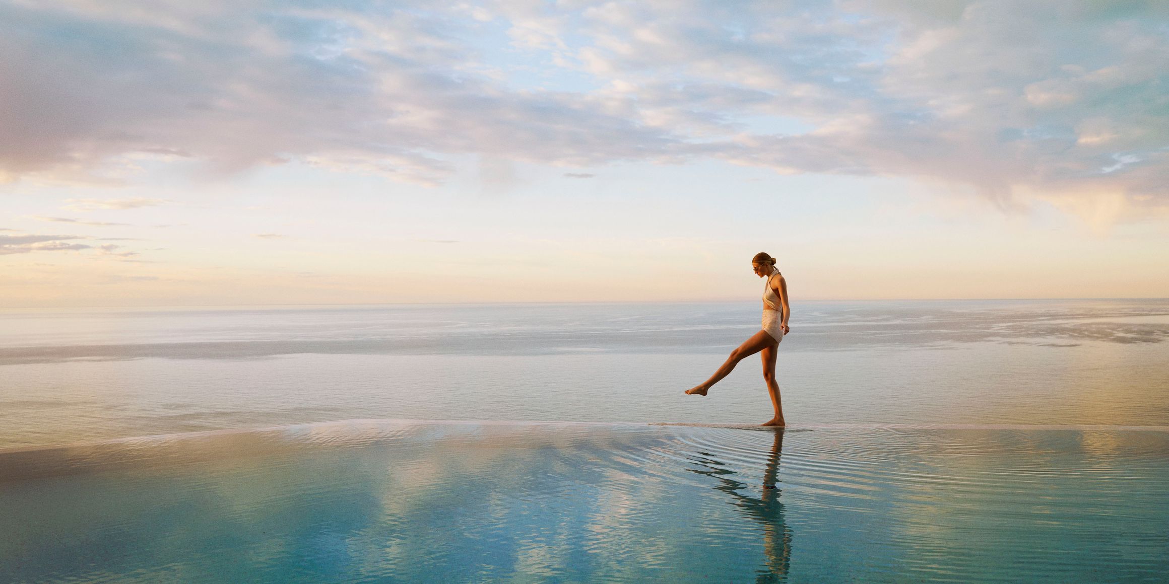 A woman in a swimsuit gracefully walks along the edge of an infinity pool, reflecting the serene ocean at sunset.