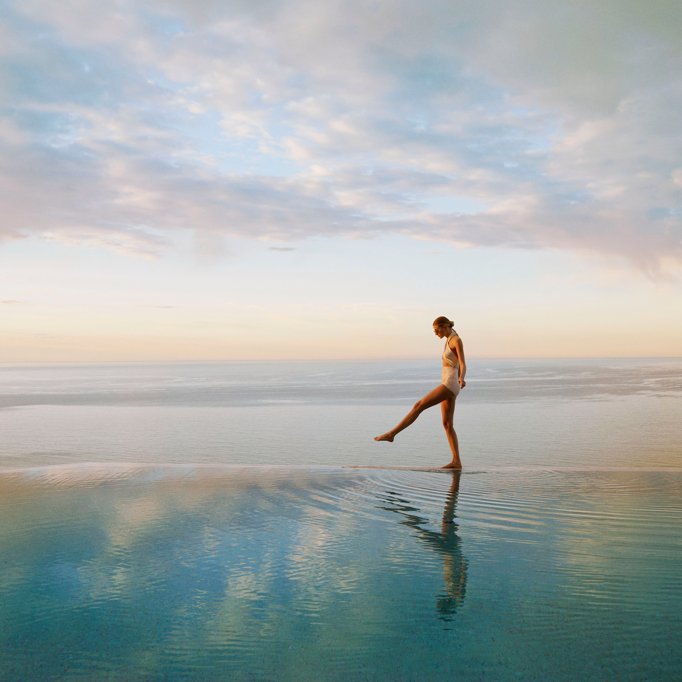 A woman in a swimsuit gracefully walks along the edge of an infinity pool, reflecting the serene ocean at sunset.