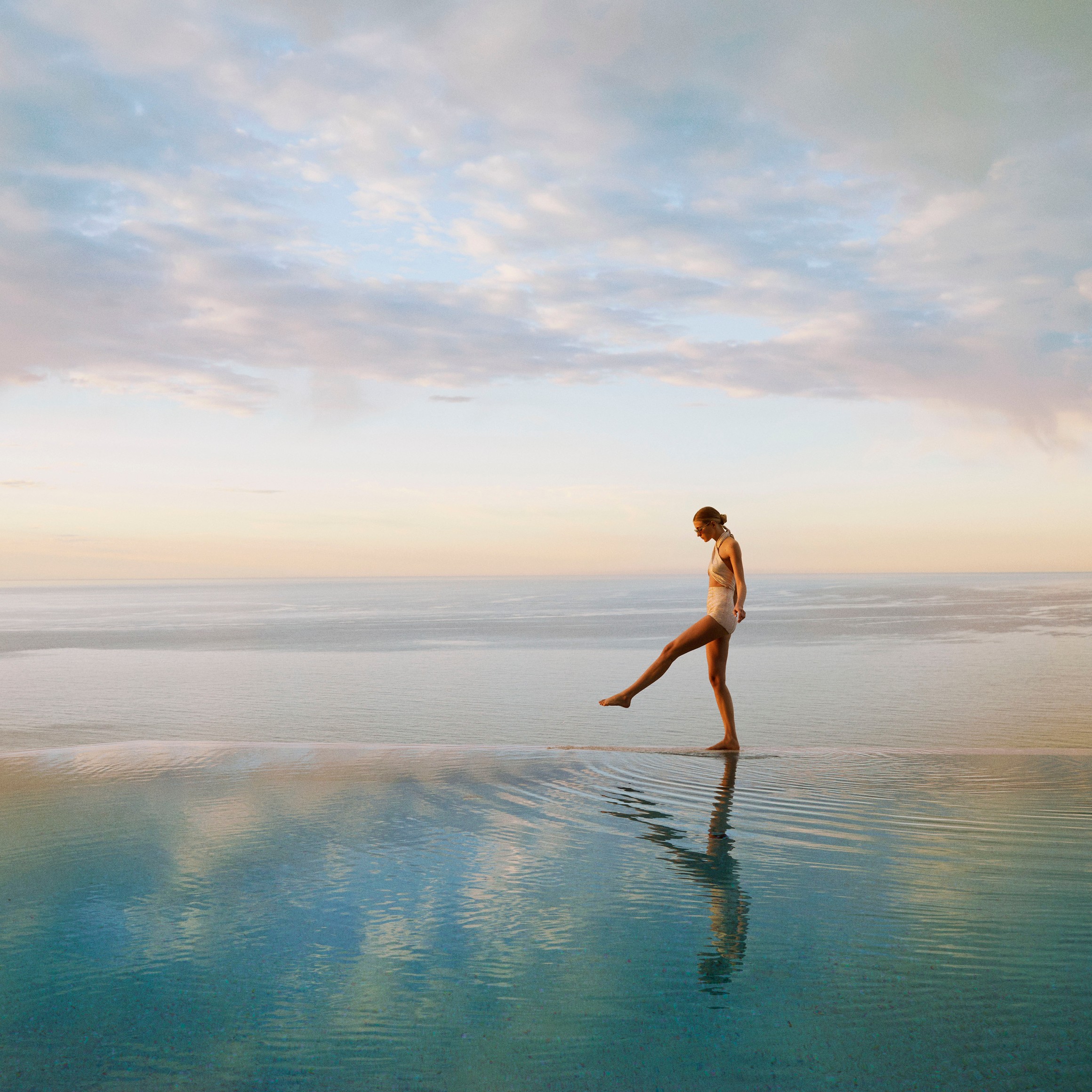 A woman in a swimsuit gracefully walks along the edge of an infinity pool, reflecting the serene ocean at sunset.