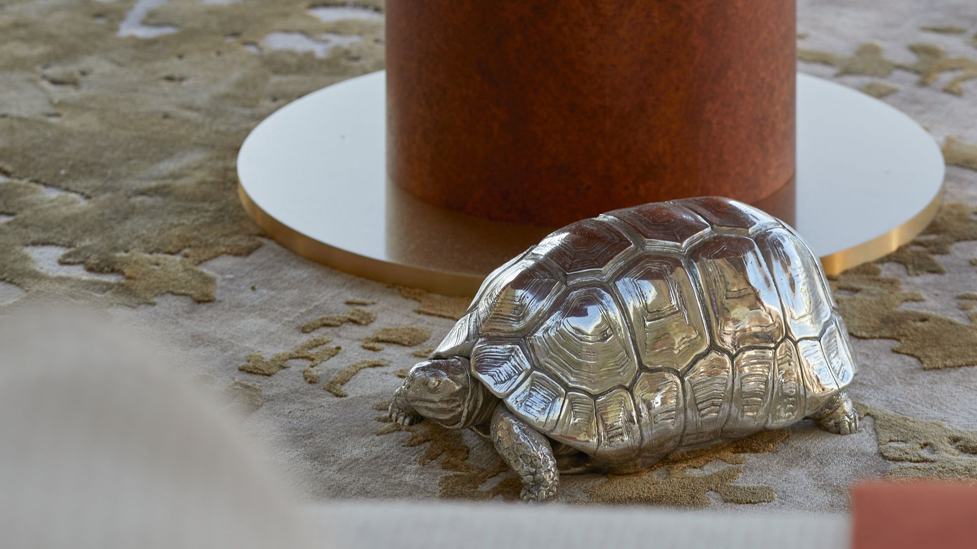 A silver tortoise figurine placed on a textured rug near a rust-hued table base, blending natural and metallic aesthetics.