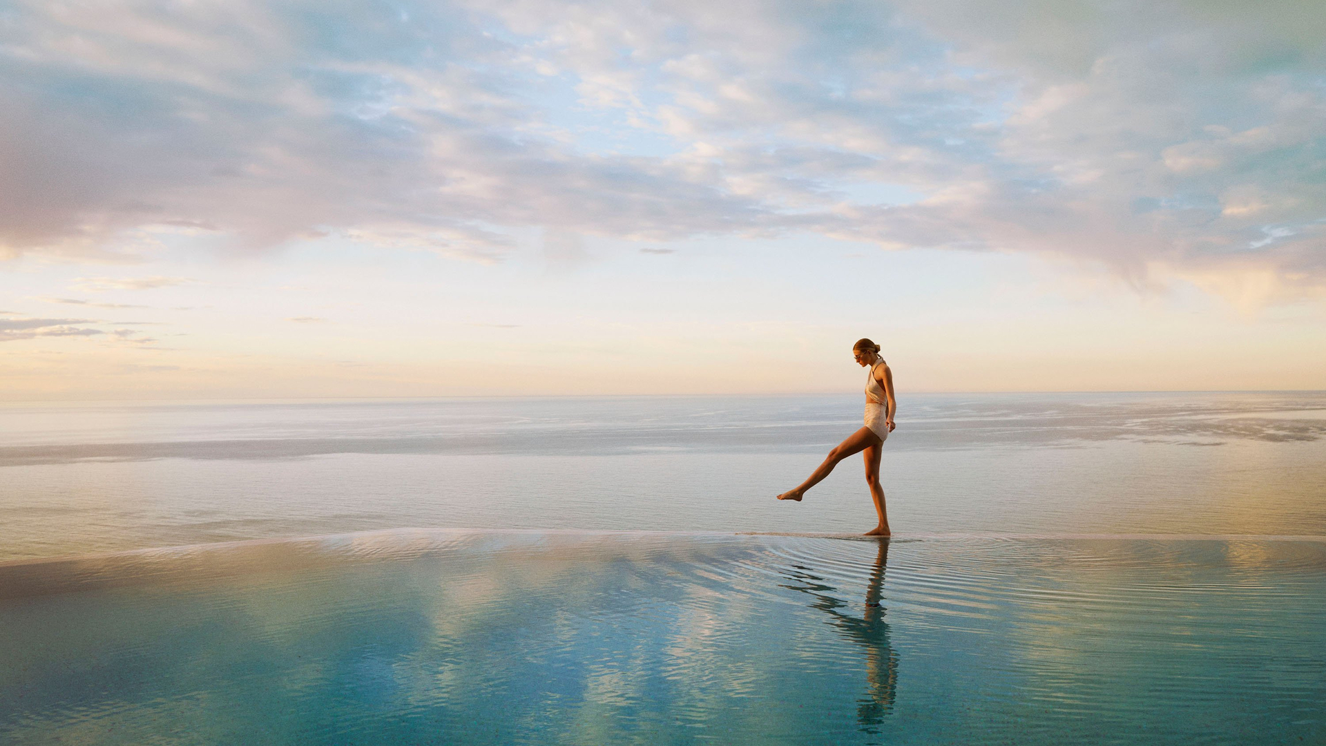 Woman in swimsuit walking along the edge of an infinity pool at sunset, overlooking a calm, expansive ocean horizon.