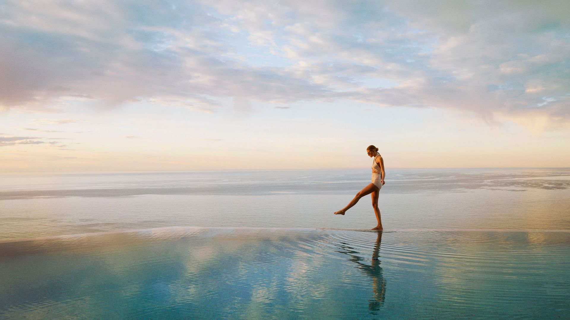 Woman in swimsuit walking along the edge of an infinity pool at sunset, overlooking a calm, expansive ocean horizon.
