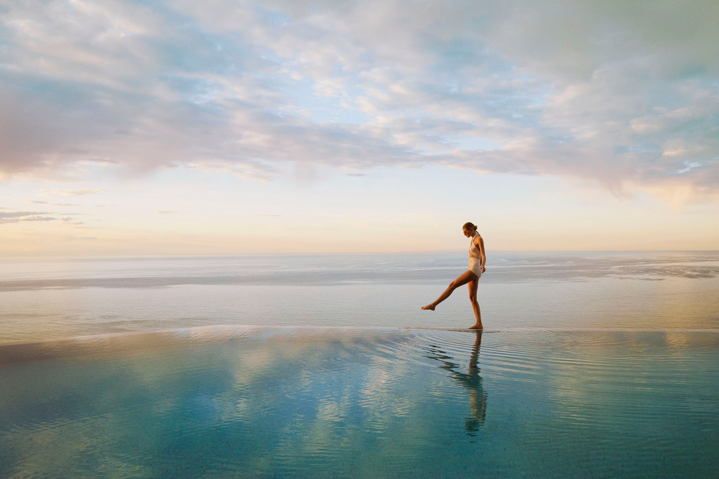 Woman in swimsuit walking along the edge of an infinity pool at sunset, overlooking a calm, expansive ocean horizon.