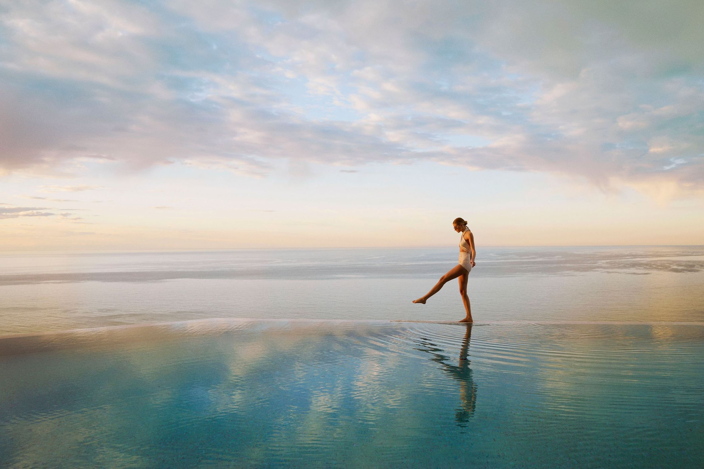 Woman in swimsuit walking along the edge of an infinity pool at sunset, overlooking a calm, expansive ocean horizon.