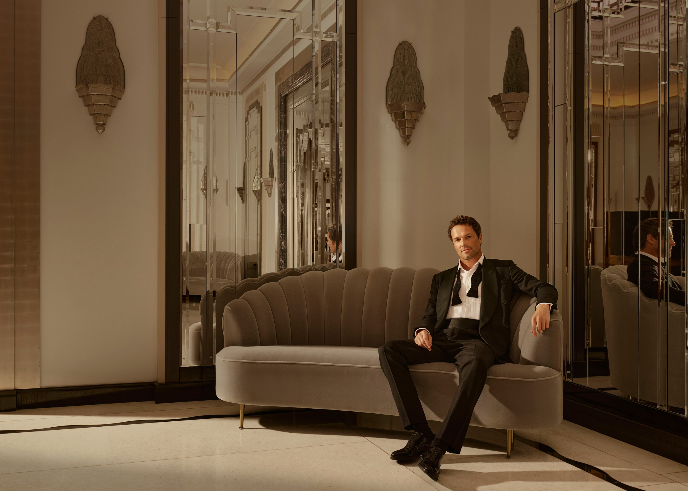 Man in a tuxedo seated on a plush sofa in an elegant, mirrored room with art deco details.